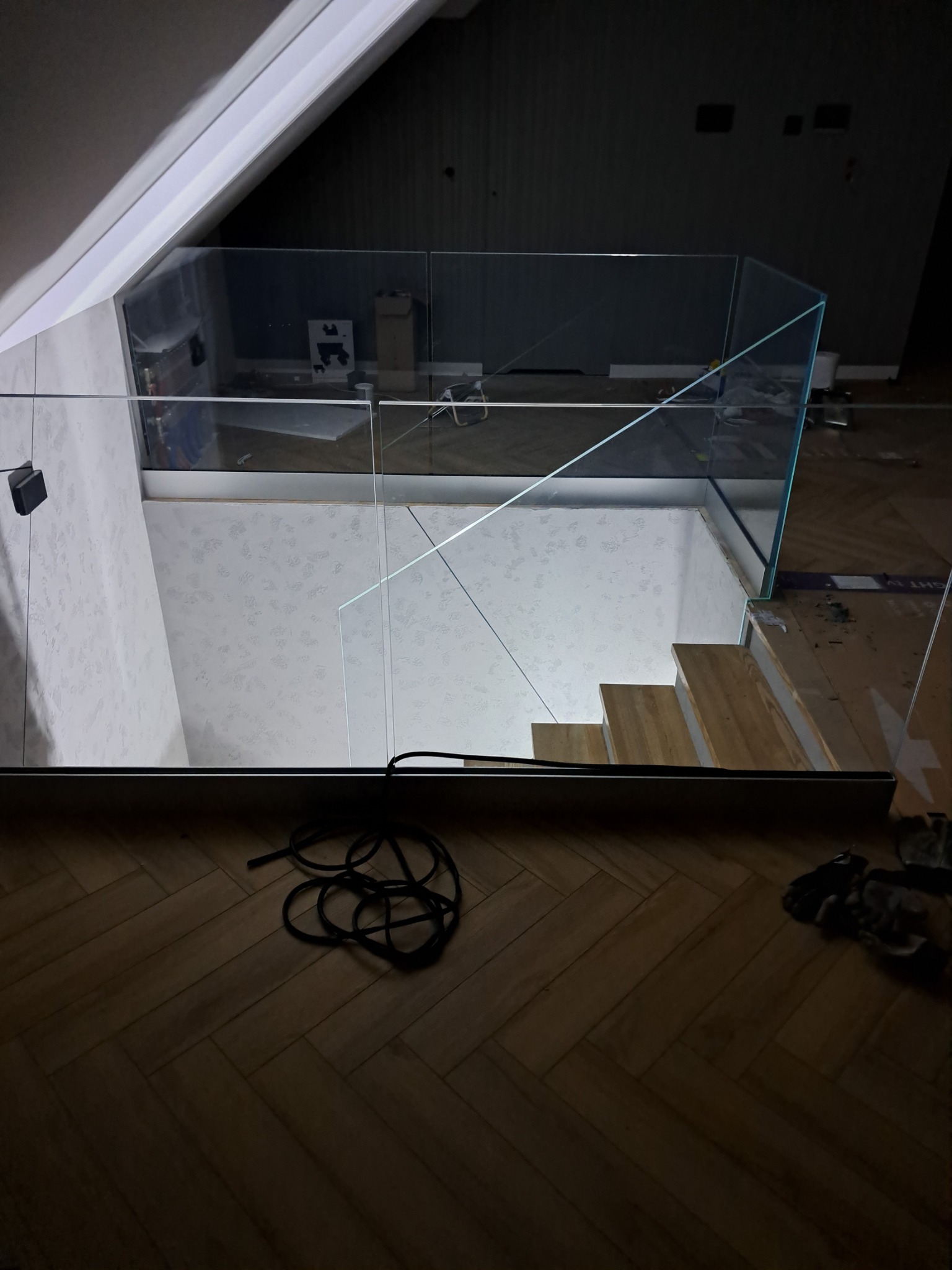 Szklana balustrada zabezpieczająca schody w domu z poddaszem, widok z góry na drewniane stopnie i fragment parkietu ułożonego w jodełkę.