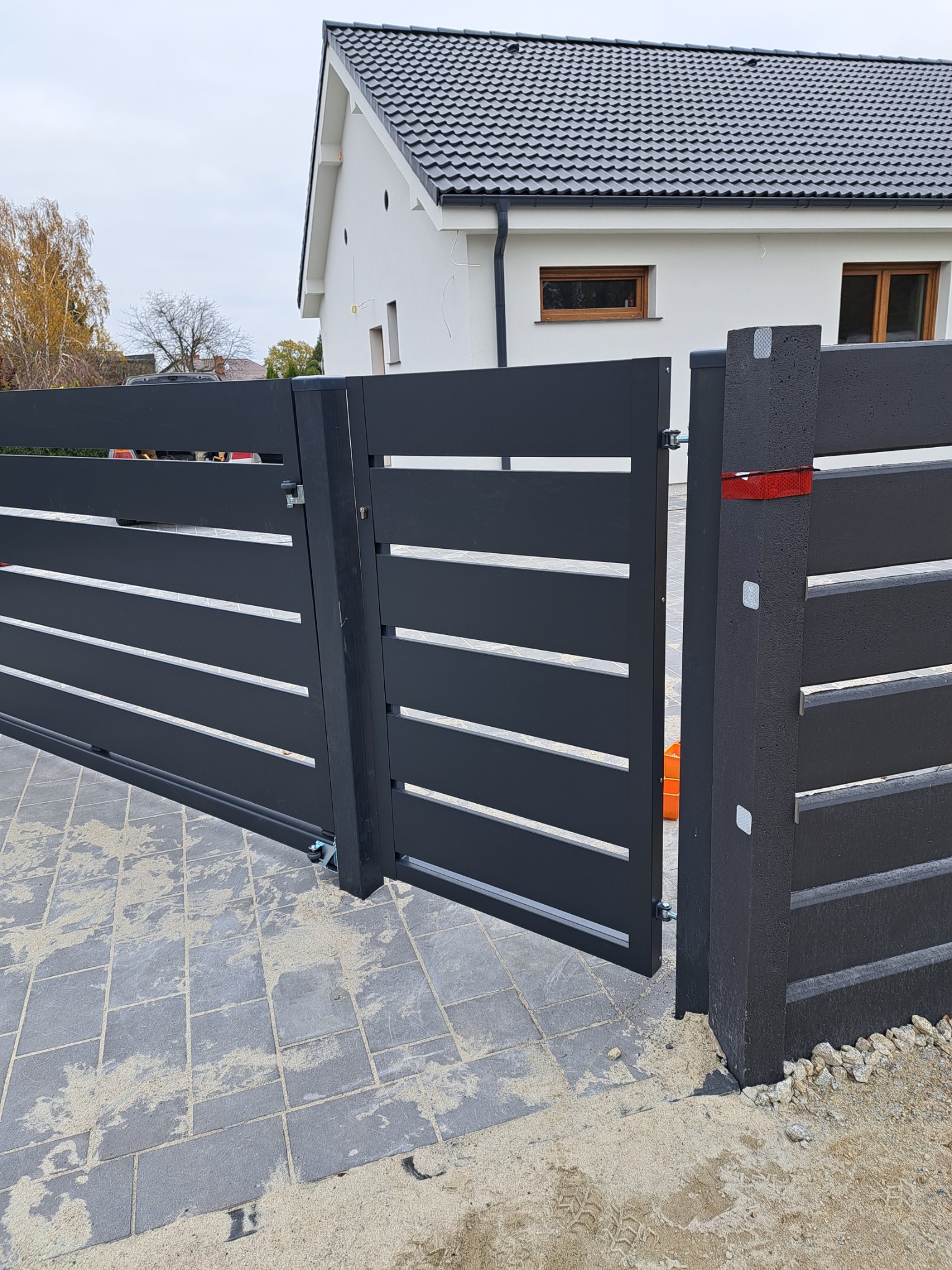 Nowoczesna, przesuwna brama wjazdowa z poziomymi panelami, pomalowana na grafitowo, z widoczną automatyką i furtką, na tle jasnego domu z ciemną dachówką i kostki brukowej.