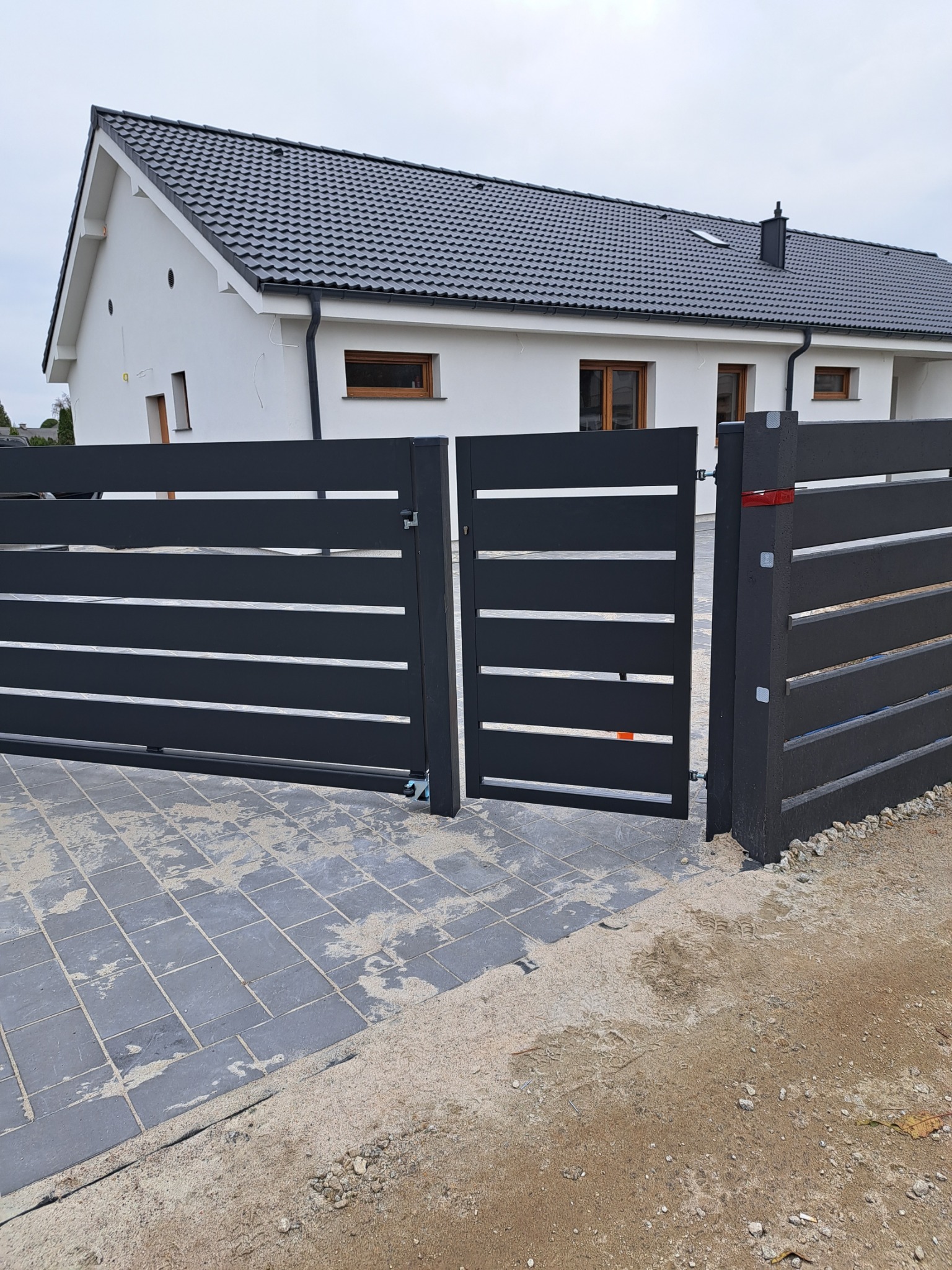 Nowoczesna, antracytowa brama wjazdowa z poziomymi panelami, częściowo otwarta, na tle białego domu z ciemną dachówką; widoczna kostka brukowa i piasek po pracach montażowych.