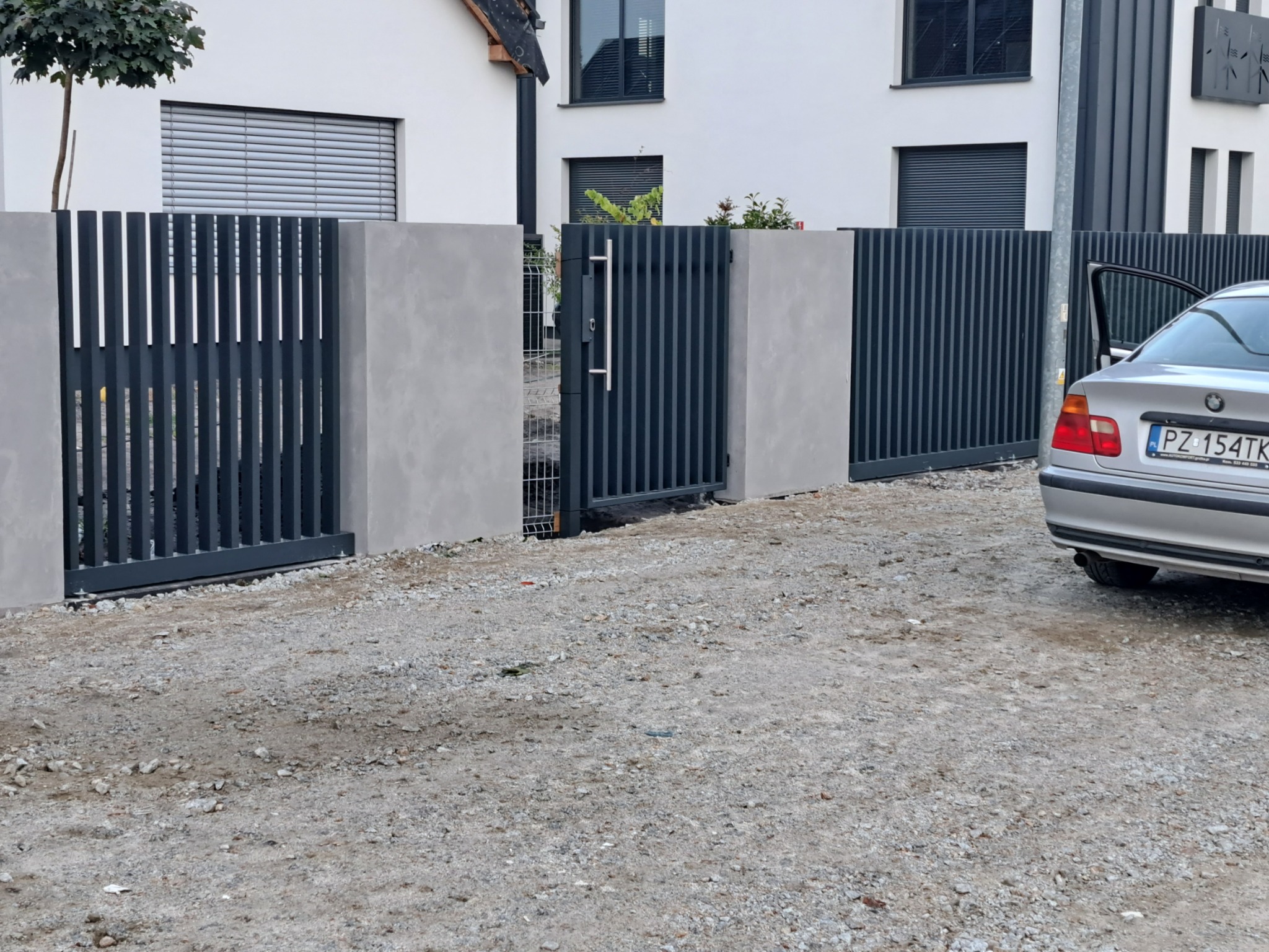Nowoczesna, antracytowa brama wjazdowa i przęsła ogrodzeniowe z pionowych profili, osadzone na betonowych słupkach, z widocznym samochodem osobowym i żwirowym podłożem.