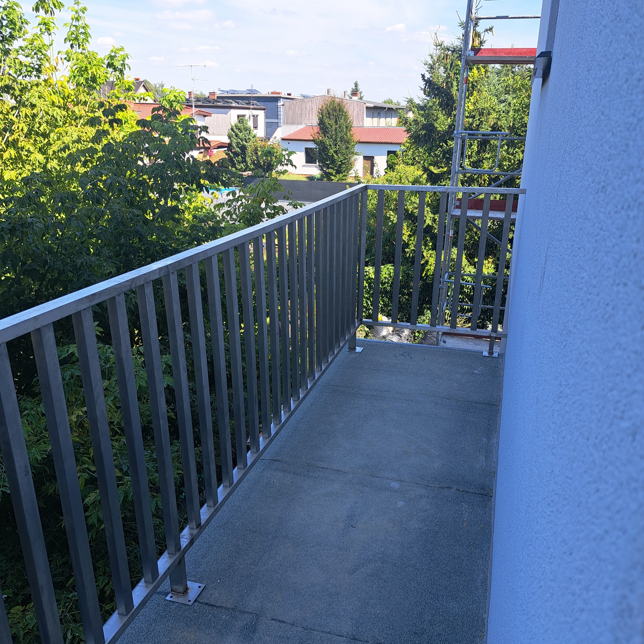 Metalowa balustrada balkonowa z widokiem na zieleń, w tle zabudowania i drabina rusztowania przy ścianie budynku.