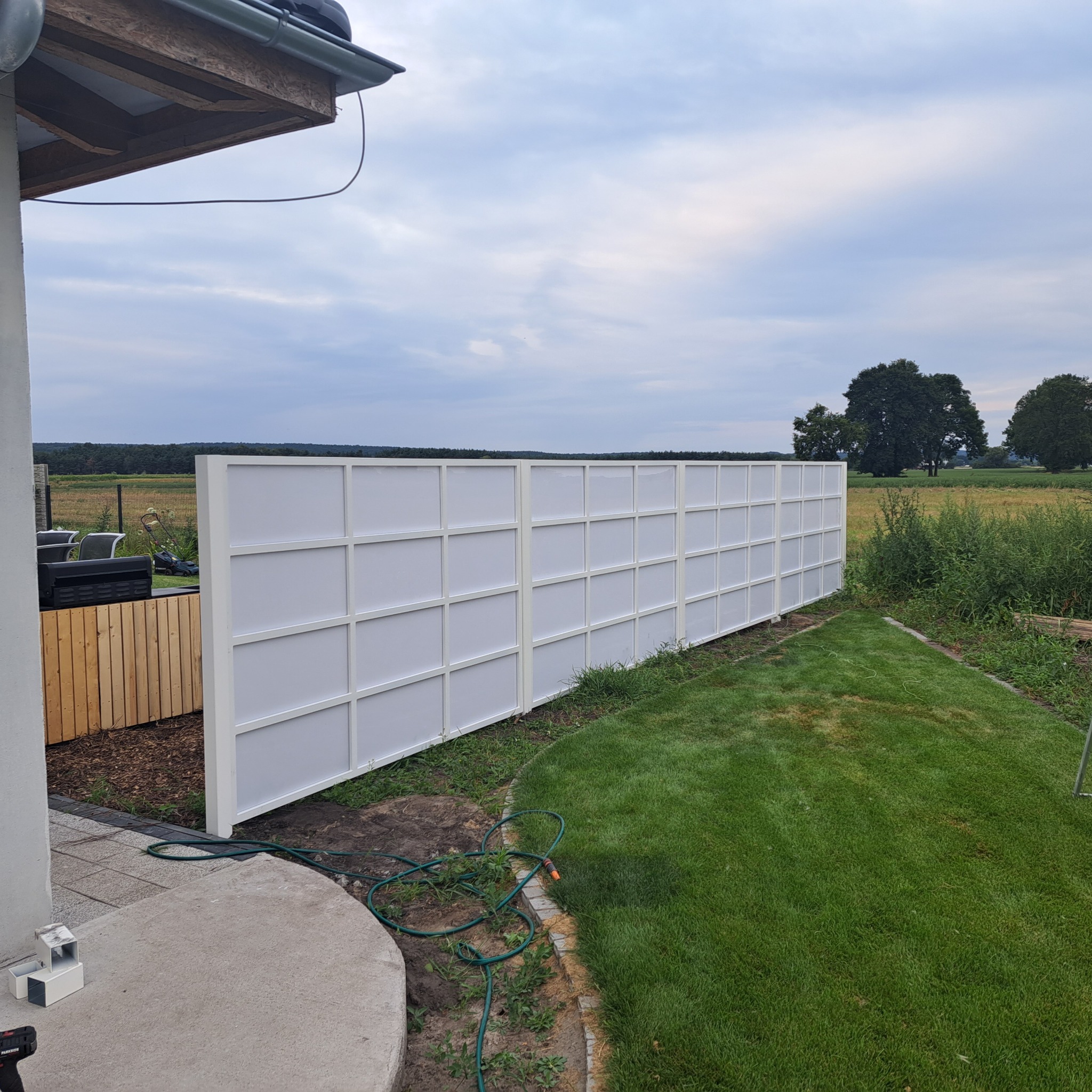 Nowoczesne białe ogrodzenie panelowe z widoczną strukturą kratową, oddzielające posesję od pola, z fragmentem tarasu i narzędziami montażowymi w kadrze.