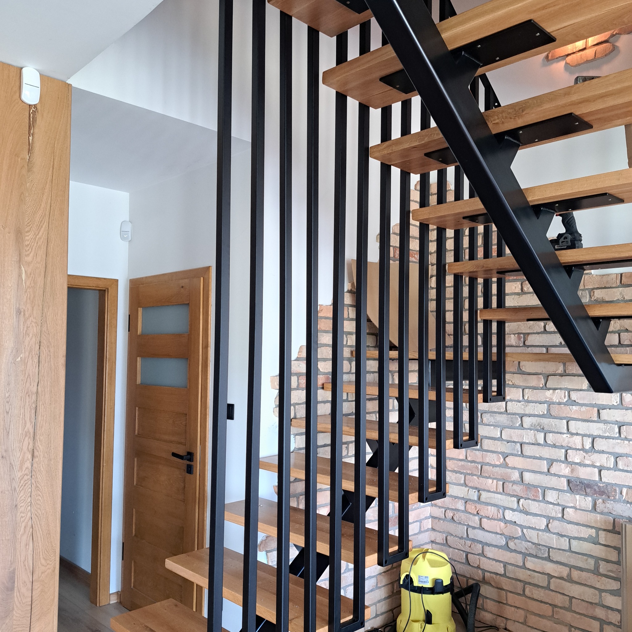 Nowoczesne schody z drewnianymi stopniami i czarną metalową balustradą na tle ceglanej ściany, widoczne drzwi wejściowe i odkurzacz.