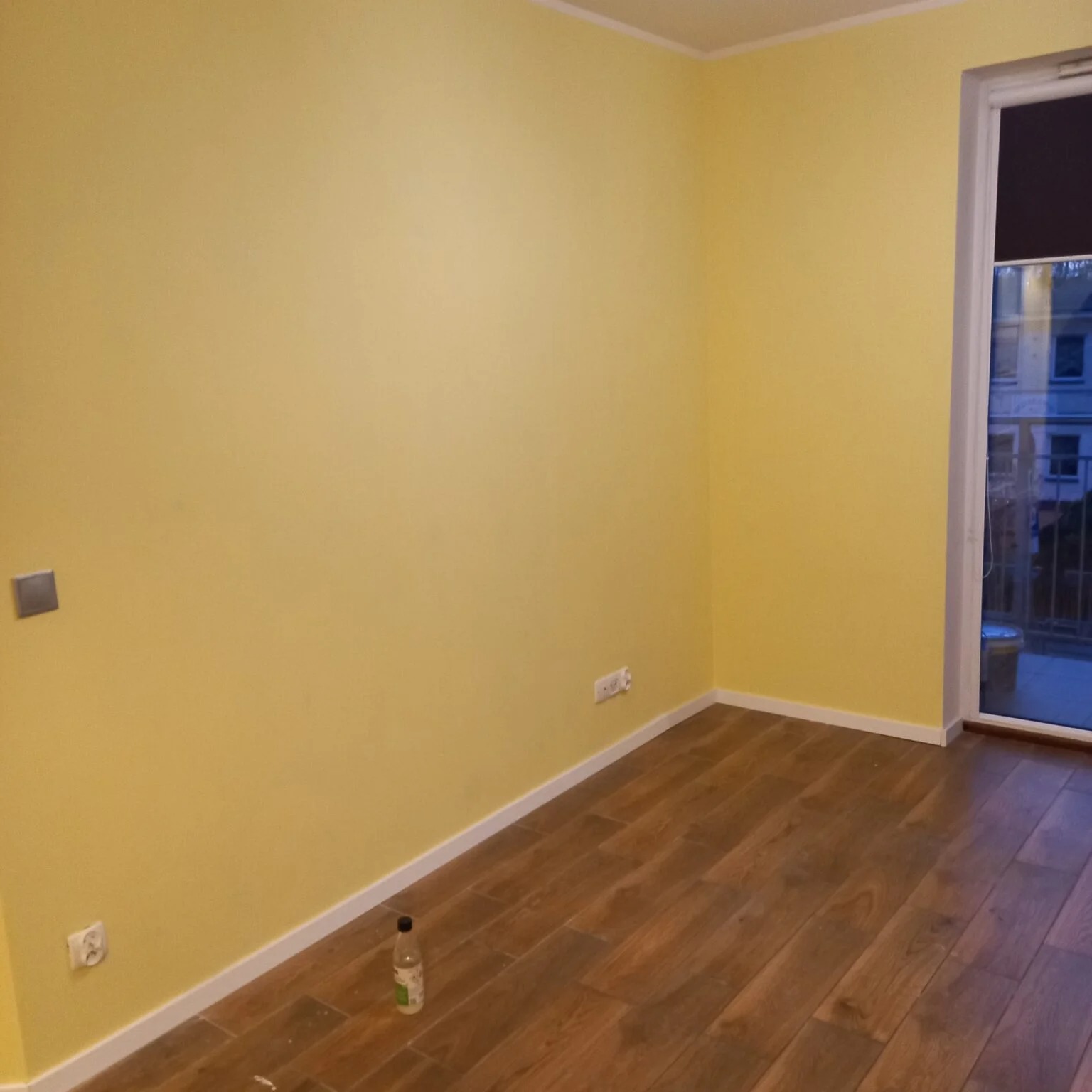 Pomalowane na żółto ściany w pokoju z brązowymi panelami podłogowymi. Widoczne gniazdka, listwy przypodłogowe i butelka na podłodze. Okno balkonowe z widokiem na zewnątrz.