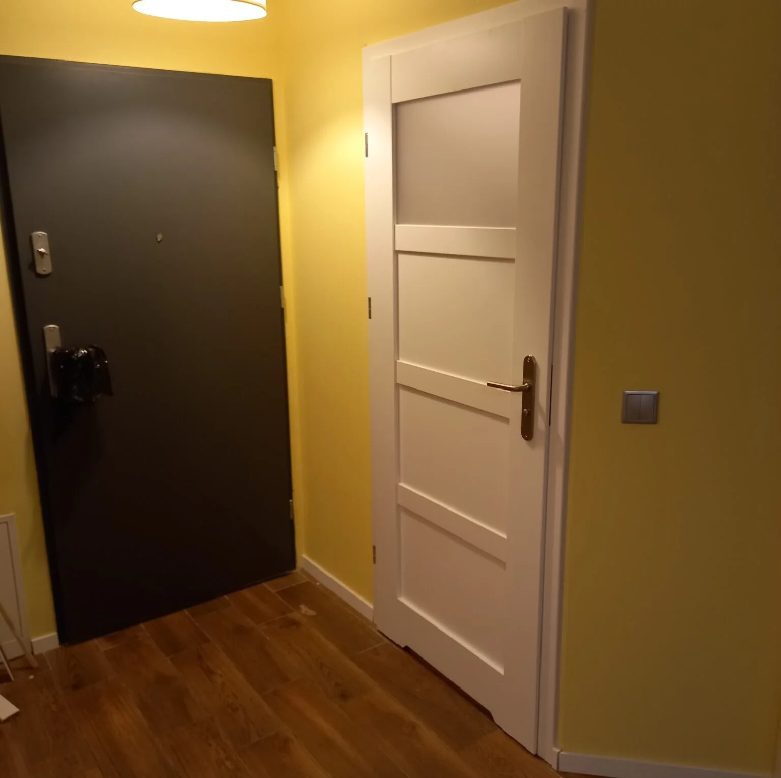 Korytarz po remoncie: żółte ściany, drewnopodobne panele, białe drzwi wewnętrzne z przeszkleniem i ciemne drzwi wejściowe. Widok na wykończone wnętrze.