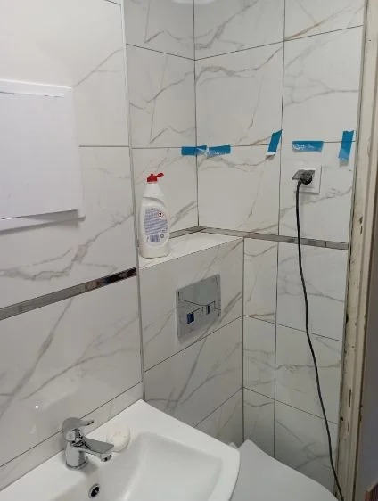 Wykończona łazienka z białymi płytkami imitującymi marmur, chromowaną armaturą i akcesoriami. Widoczne niebieskie taśmy maskujące na fugach.