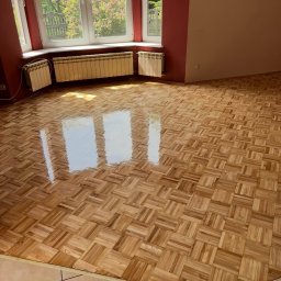 Aleksander Waliczek - Parkiet po renowacji w jasnym pokoju z oknem i grzejnikami. Widoczne odbicie światła na gładkiej powierzchni podłogi. Ściany w odcieniach czerwieni i beżu.