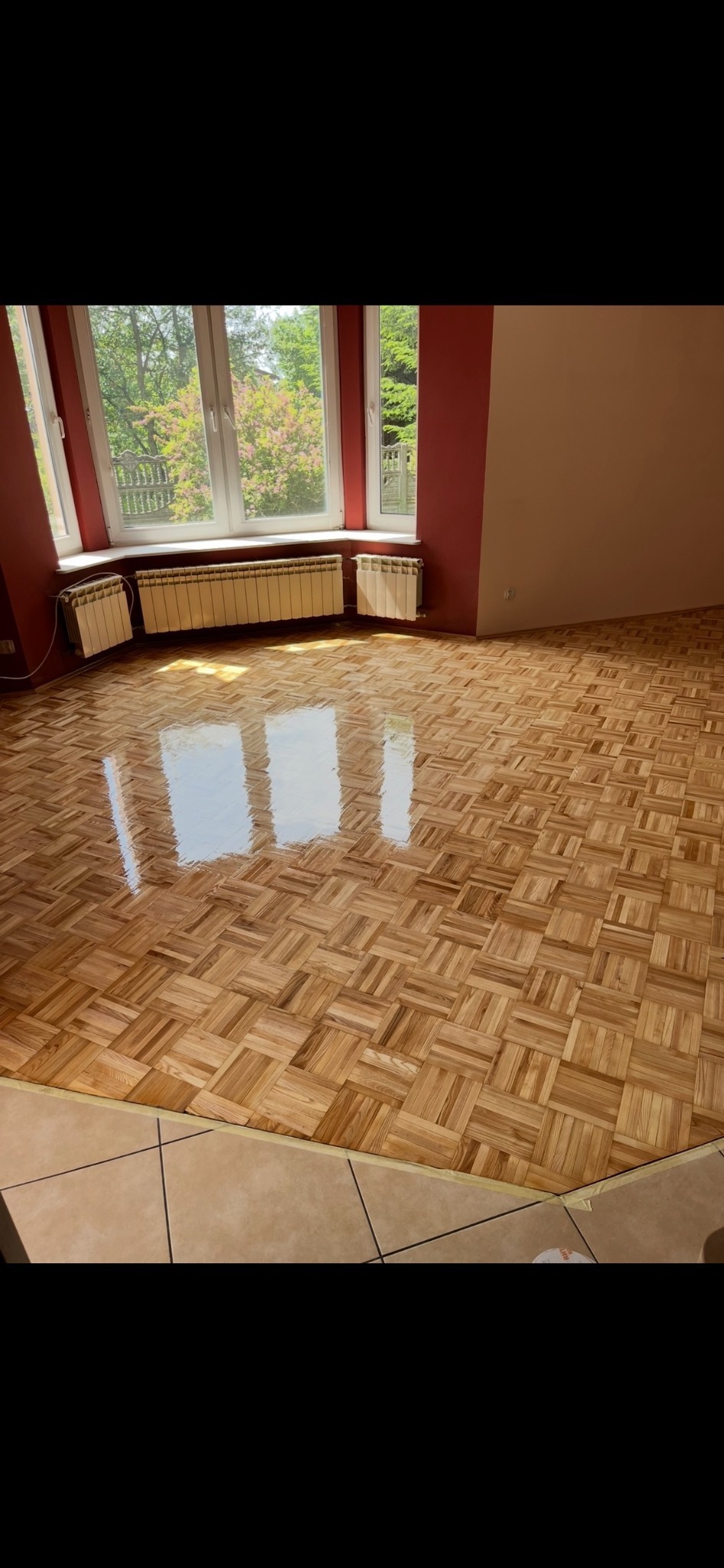 Parkiet po renowacji w jasnym pokoju z oknem i grzejnikami. Widoczne odbicie światła na gładkiej powierzchni podłogi. Ściany w odcieniach czerwieni i beżu.
