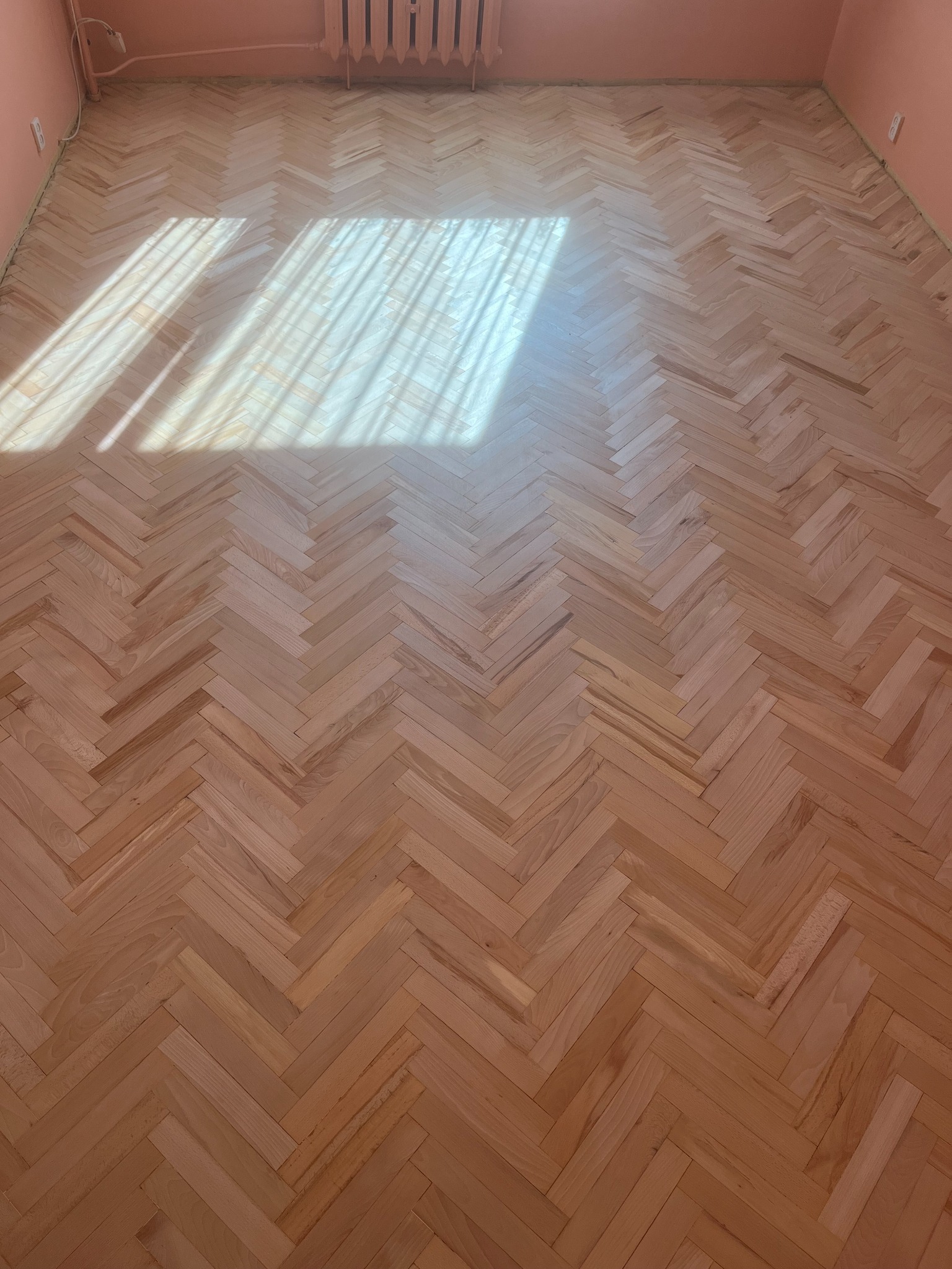 Jasna, odnowiona podłoga parkietowa ułożona w jodełkę, oświetlona promieniami słońca wpadającymi przez okno. Widoczne ślady po szlifowaniu.