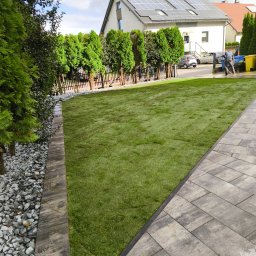TRYBA OGRODY - Świeżo ułożona trawa z rolki w ogrodzie, widok z poziomu chodnika. Granica trawnika wykończona kamieniami i obrzeżem. W tle dom z panelami słonecznymi i osoba myjąca chodnik.