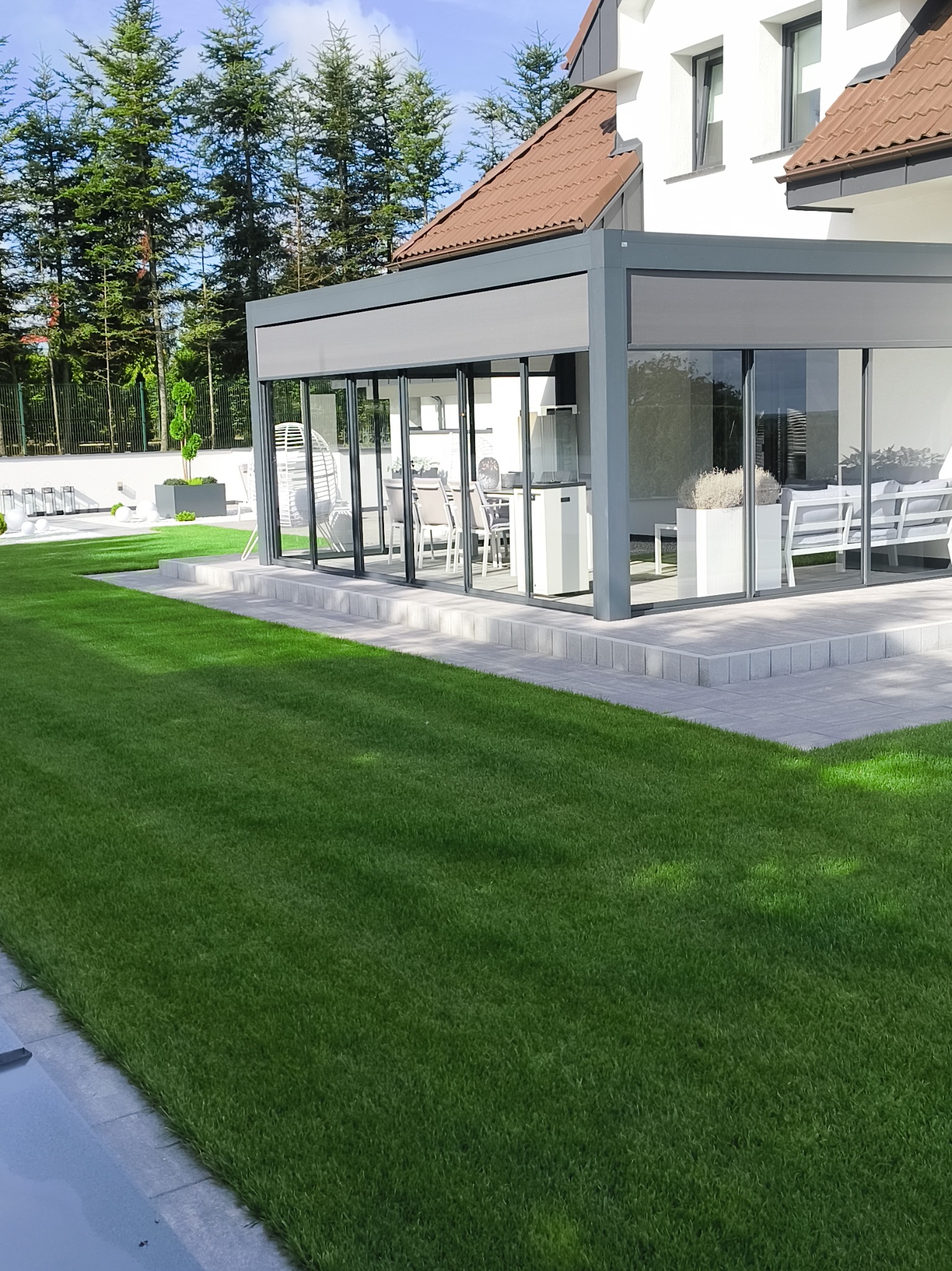 Zadbany trawnik przed nowoczesną, przeszkloną altaną z szarymi ramami. W tle wysokie drzewa iglaste i biały budynek z brązowym dachem. Widoczne elementy dekoracyjne ogrodu.
