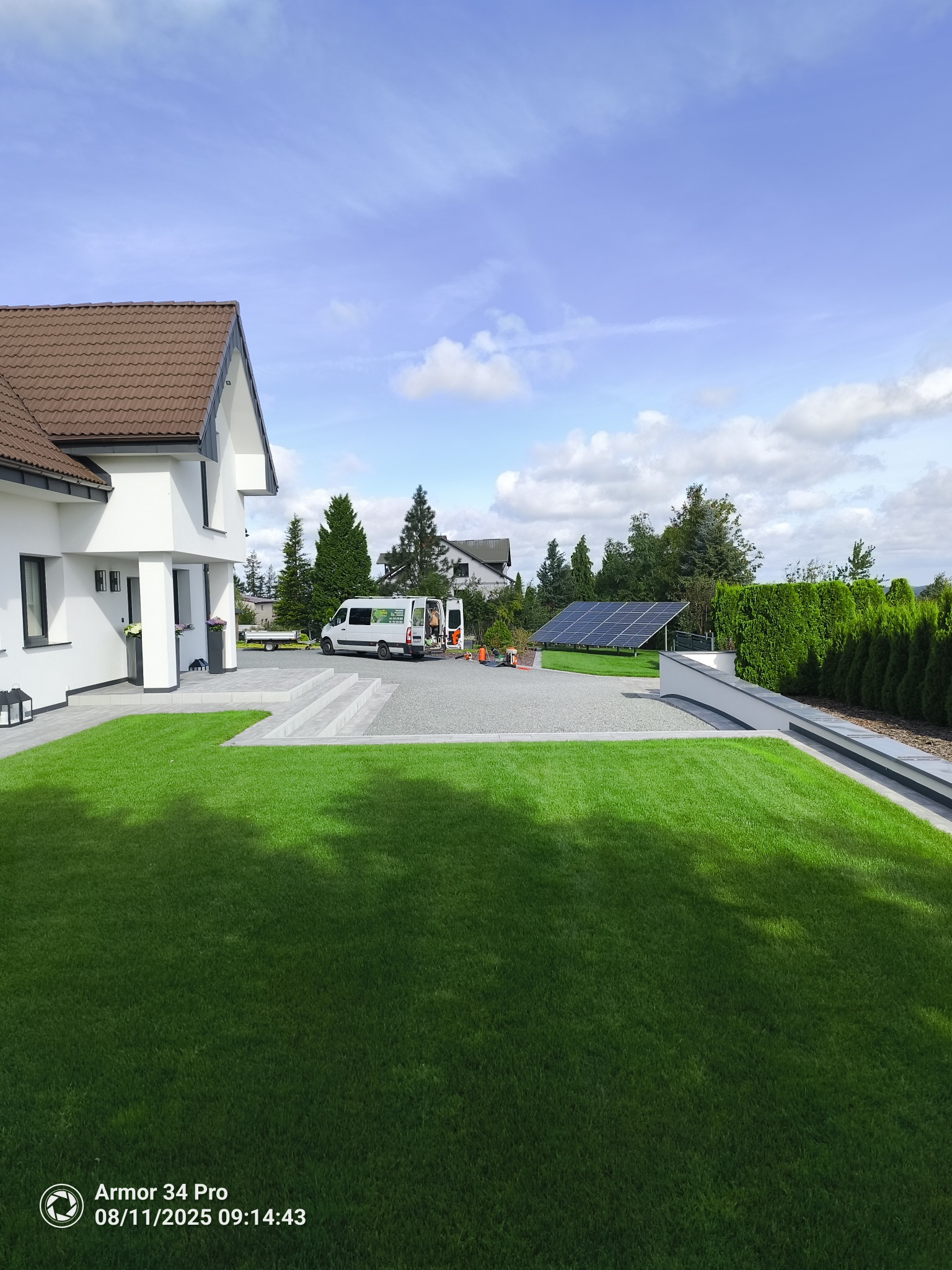Nowoczesny dom z zadbanym trawnikiem, szarym podjazdem i panelem słonecznym w tle. Na podjeździe stoi bus firmy ogrodniczej z przyczepką.