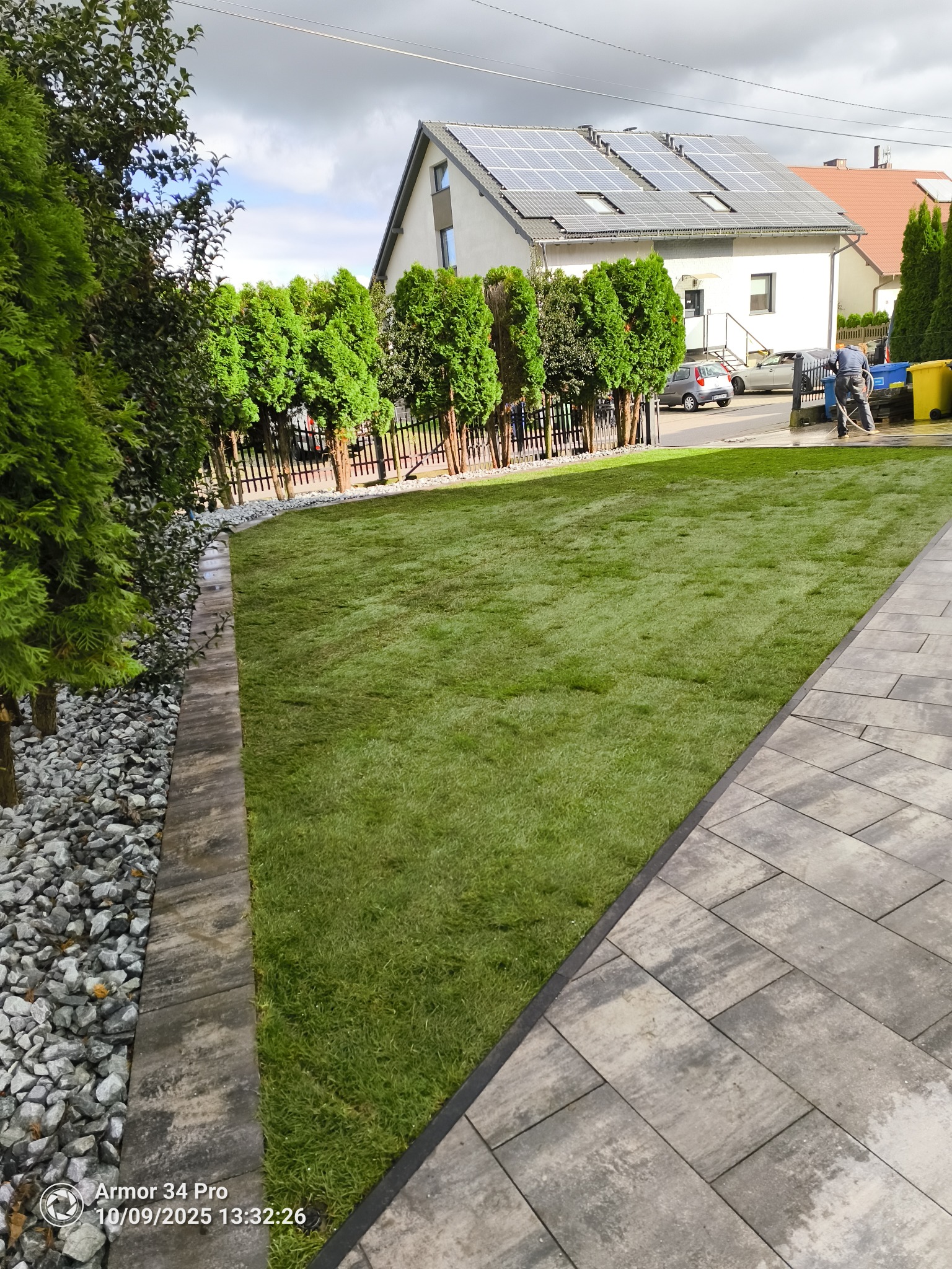 Świeżo ułożona trawa z rolki w ogrodzie, widok z poziomu chodnika. Granica trawnika wykończona kamieniami i obrzeżem. W tle dom z panelami słonecznymi i osoba myjąca chodnik.