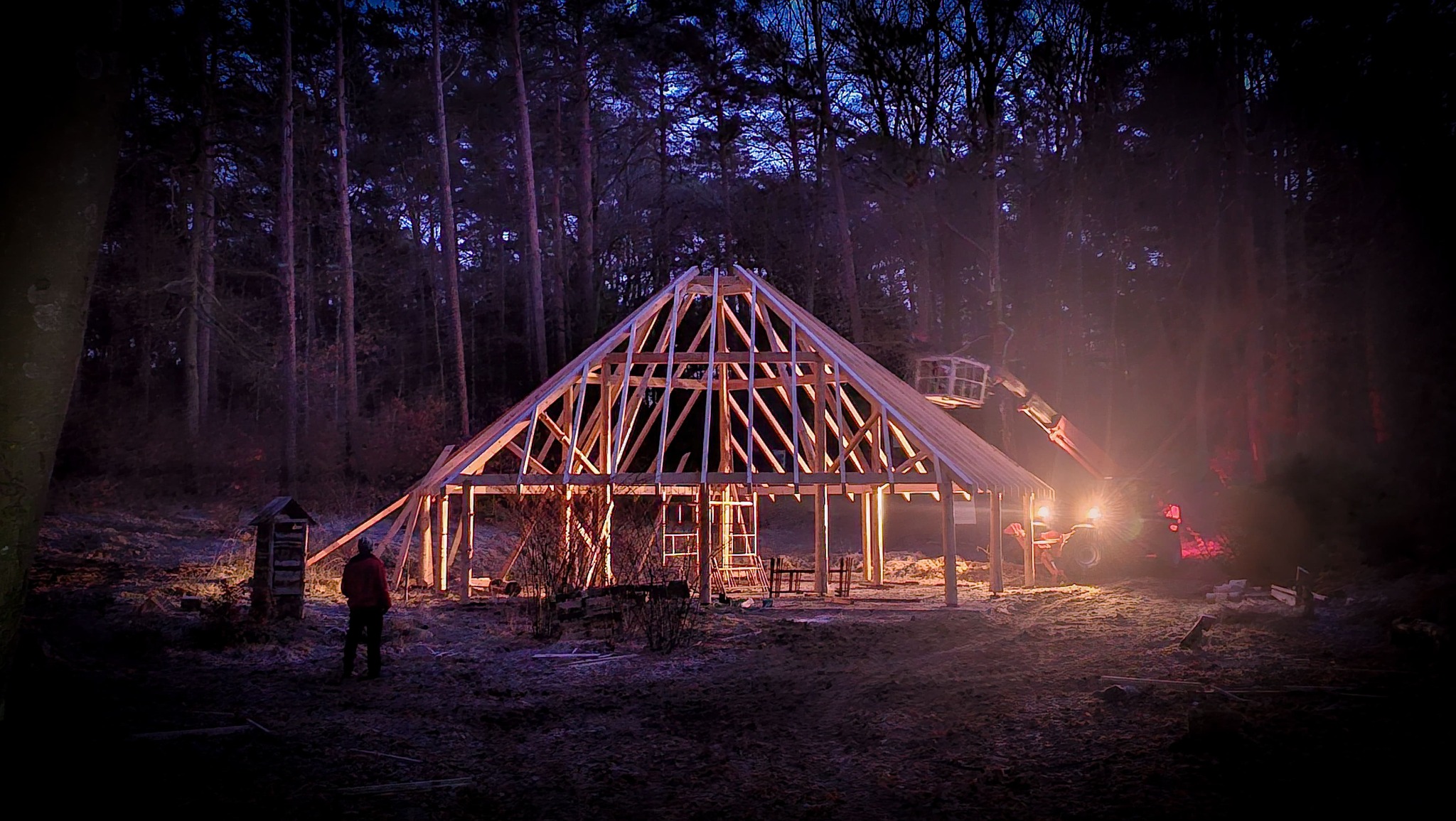 Wznoszenie drewnianej konstrukcji z więźbą dachową w leśnej scenerii, oświetlonej światłami budowlanymi i z widocznym podnośnikiem teleskopowym. Osoba obserwuje postęp prac.