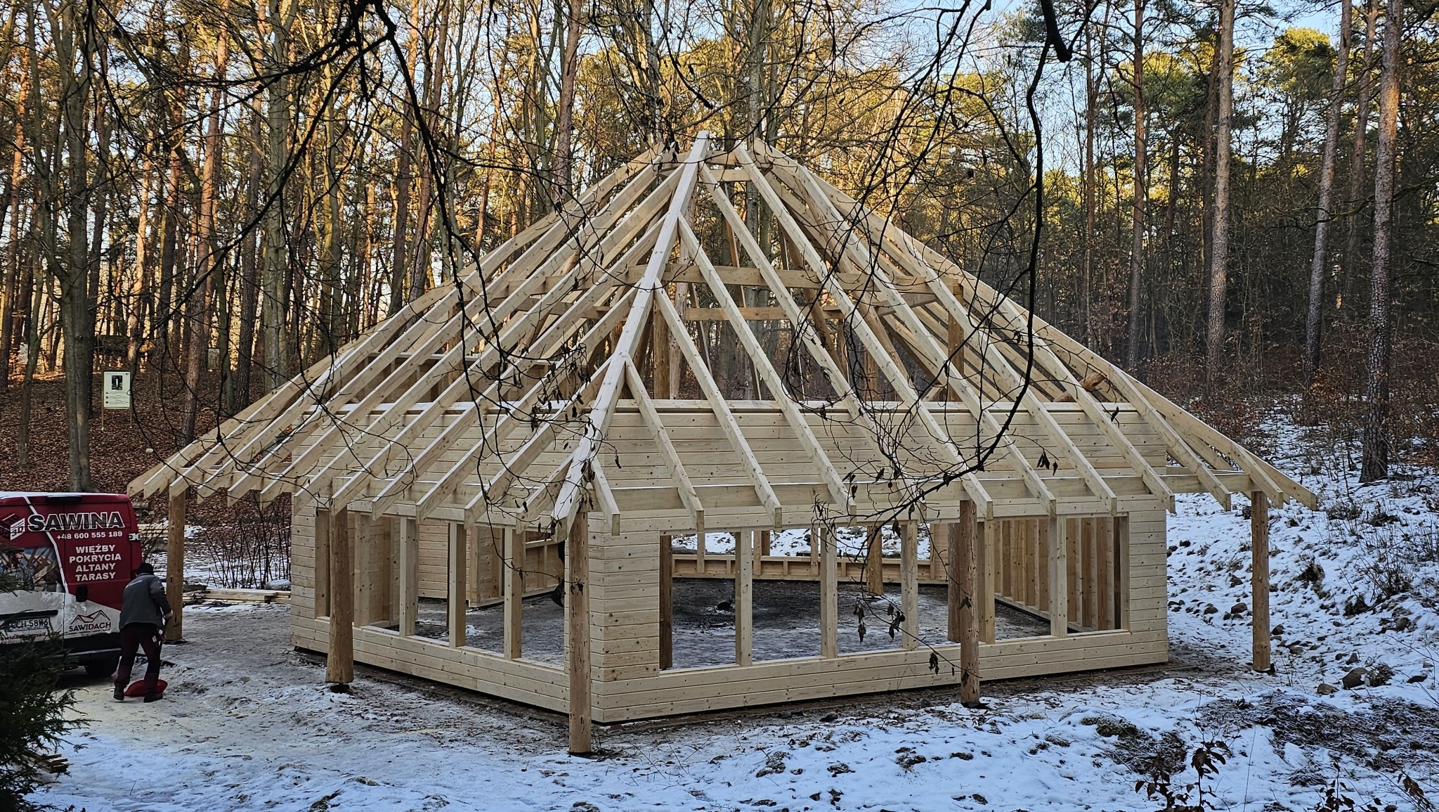 Surowa konstrukcja drewnianej altany z widoczną więźbą dachową na tle zimowego lasu. Na pierwszym planie częściowo widoczny samochód dostawczy.