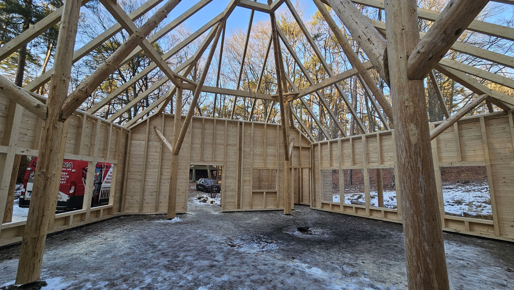 Surowa konstrukcja drewniana budynku w lesie, widok od wewnątrz. Ściany z desek, otwory okienne i drzwiowe, widoczna więźba dachowa. Częściowo ośnieżone podłoże.