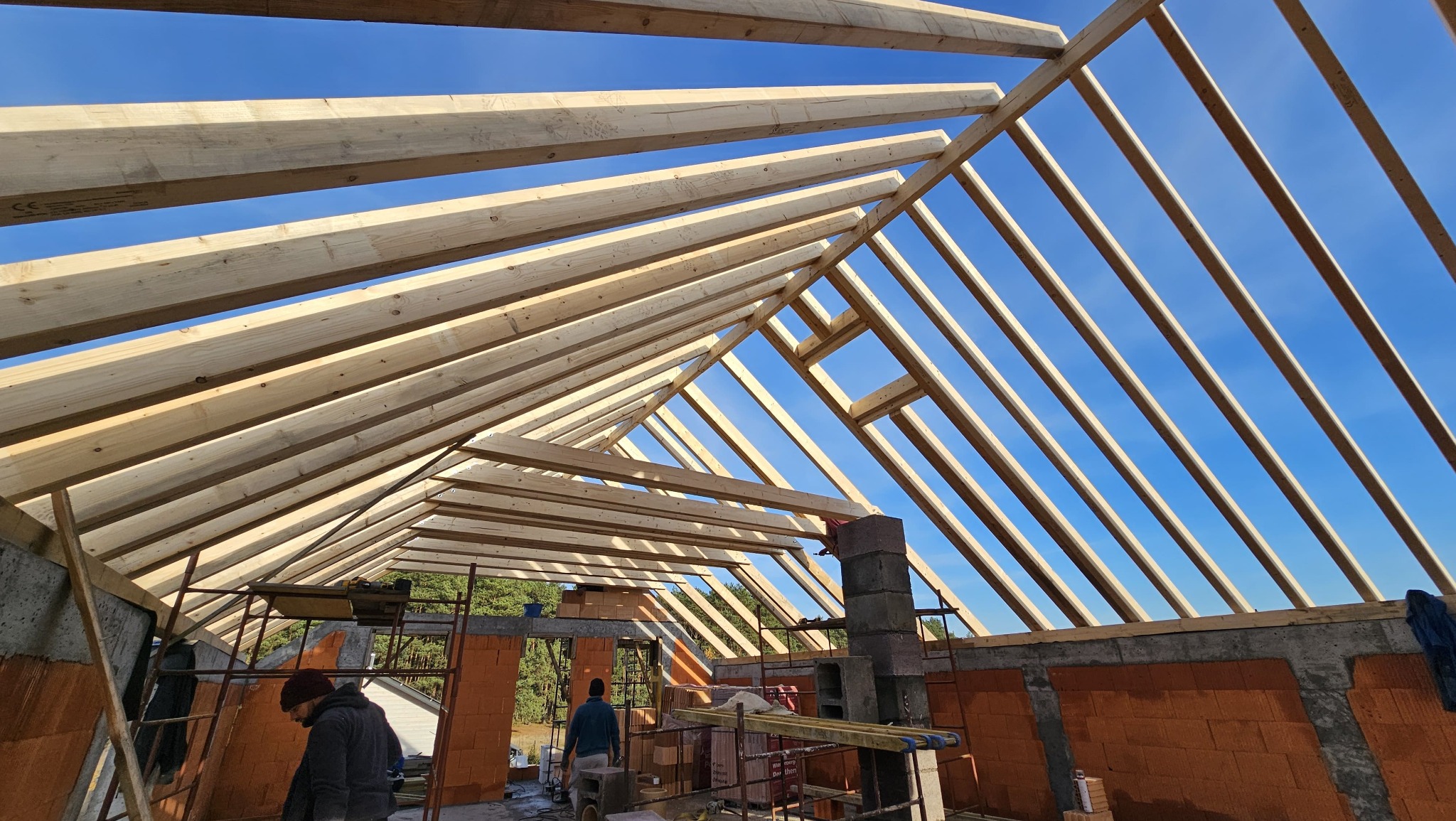 Wnętrze budowy domu z widoczną drewnianą konstrukcją dachu na tle błękitnego nieba. Widoczne ceglane ściany i rusztowania. Dwóch pracowników w tle.