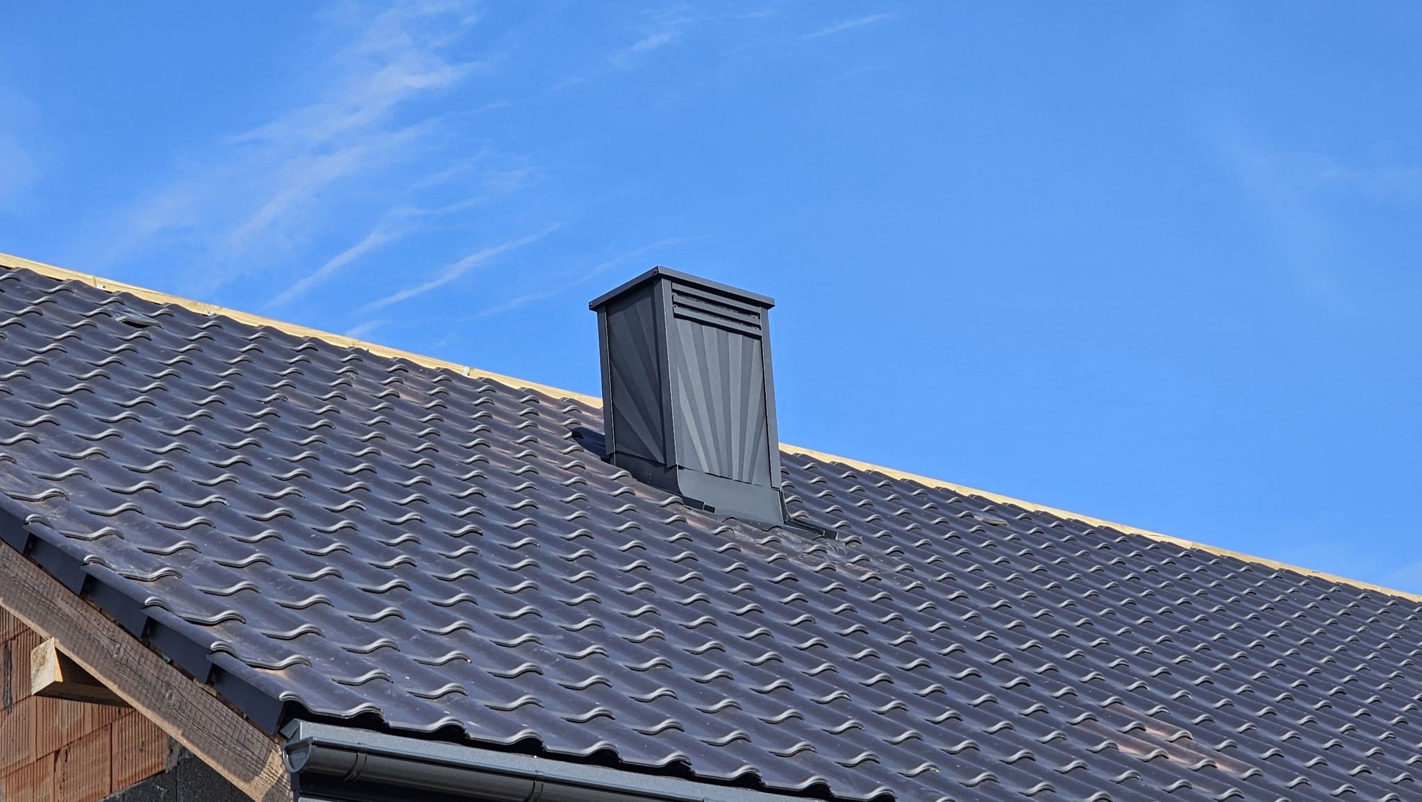 Ciemna dachówka ceramiczna na dachu domu w słoneczny dzień. Widoczny komin z blachy w kolorze dachówki i fragment orynnowania. Dach w trakcie budowy.