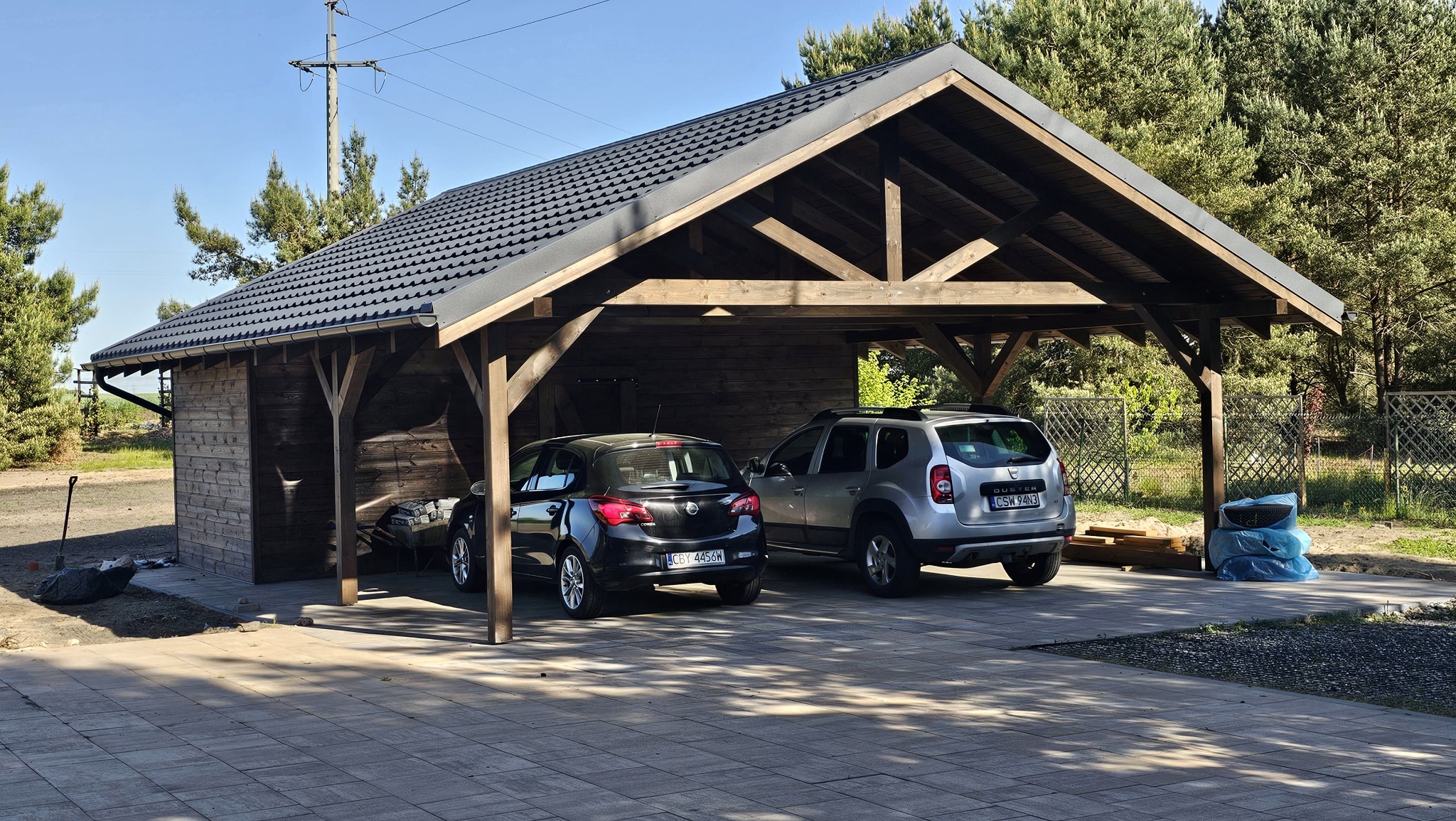 Drewniana wiata garażowa z dwoma samochodami, kryta ciemną dachówką, z widoczną konstrukcją więźby. Ujęcie w słoneczny dzień, z elementami ogrodu w tle.