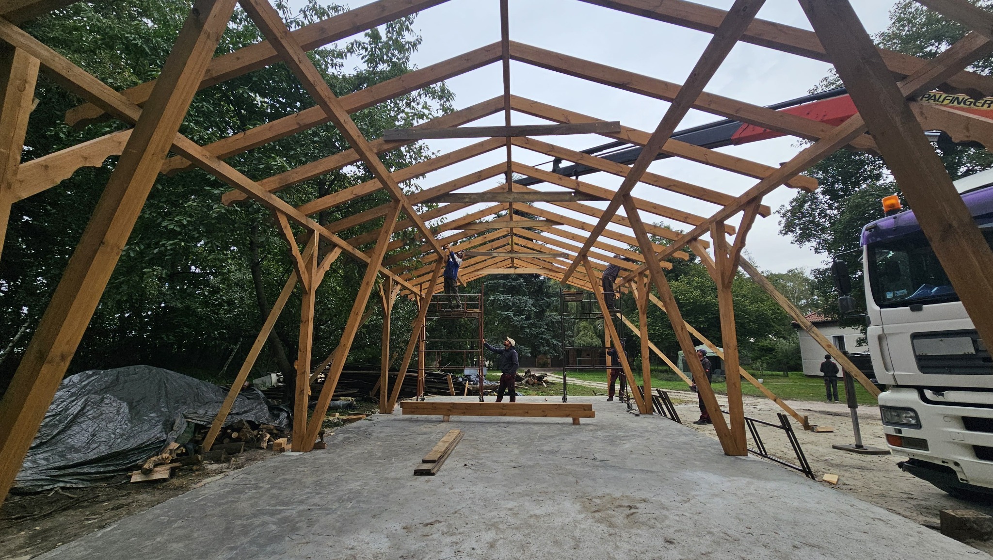 Montaż drewnianej konstrukcji z użyciem dźwigu. Widoczni pracownicy na rusztowaniach. Elementy dekoracyjne na końcach belek. Częściowo widoczny samochód ciężarowy.