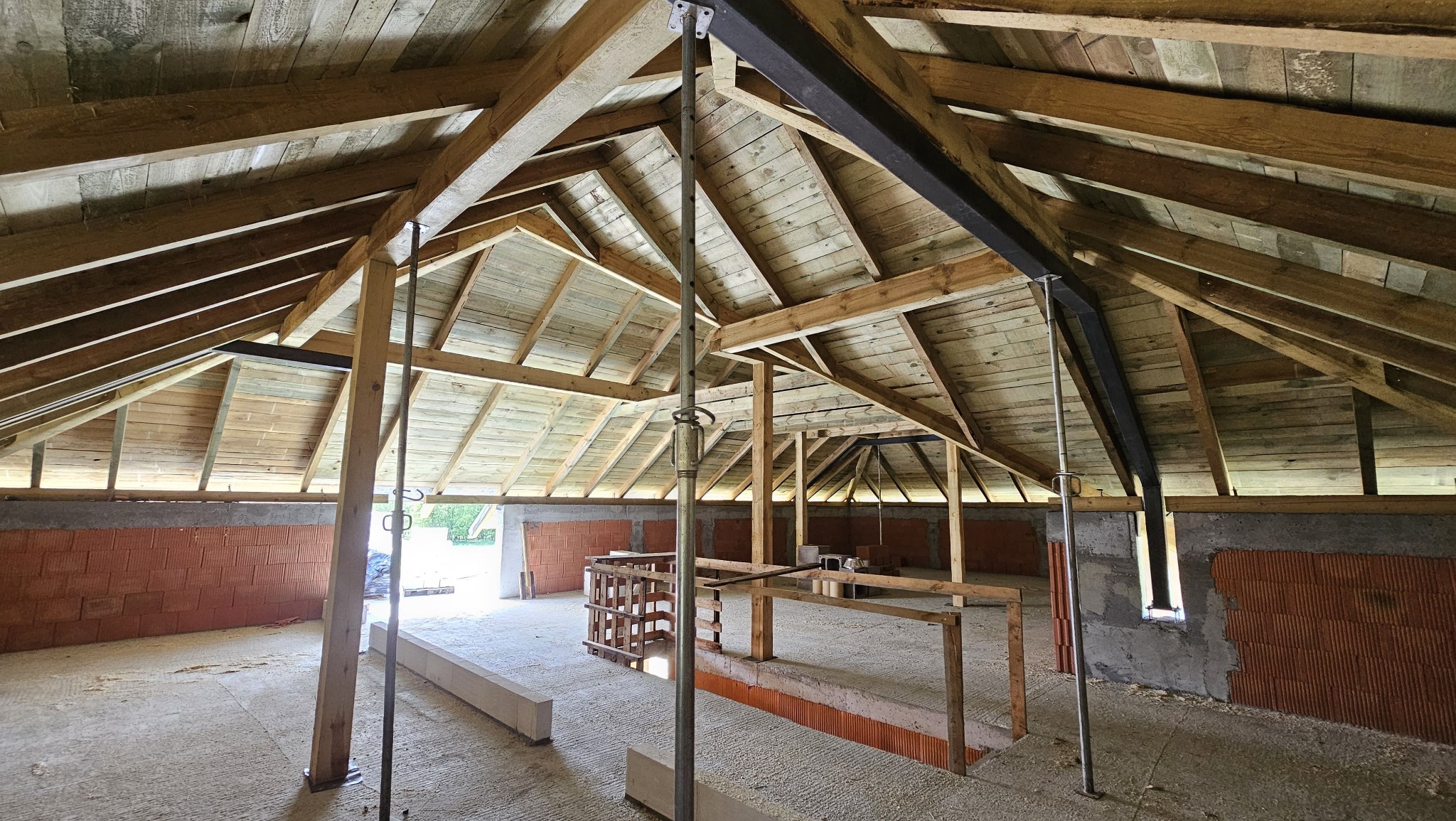 Wnętrze w trakcie budowy z widoczną drewnianą konstrukcją dachu, wspartą metalowymi stemplami. Ściany z czerwonej cegły dziurawki. Surowy stan wykończenia.