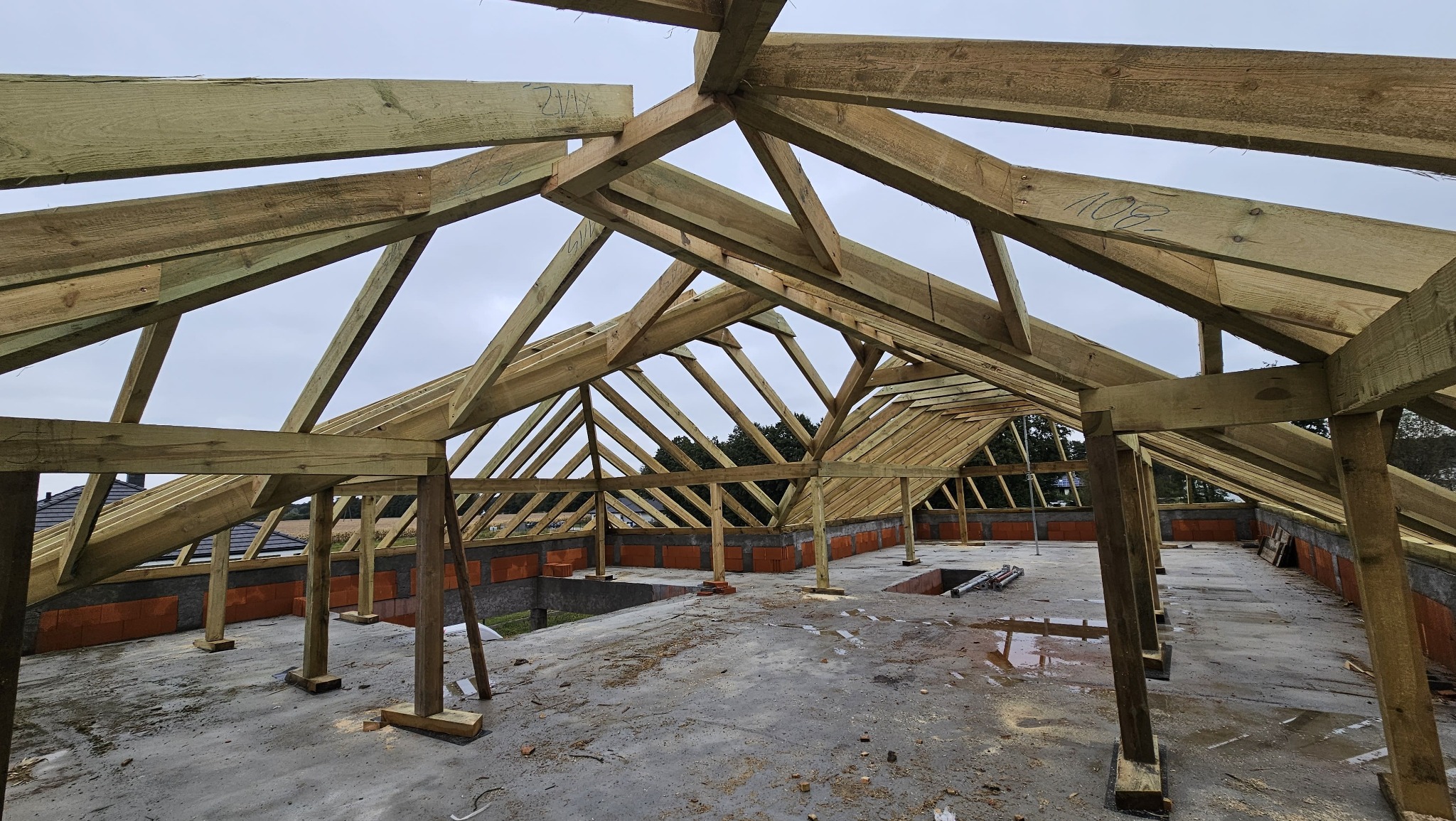 Wnętrze budynku w trakcie budowy z widoczną drewnianą konstrukcją więźby dachowej, ceglanymi ścianami i betonową podłogą. Widoczne oznaczenia na elementach drewnianych.
