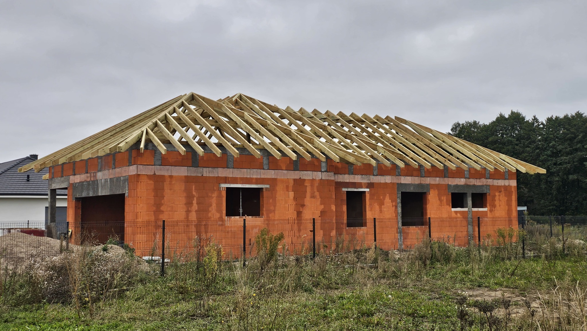 Surowy stan domu z czerwonej cegły z drewnianą konstrukcją dachu w trakcie budowy, ogrodzony siatką. Widoczne okna i garaż. W tle zieleń i pochmurne niebo.