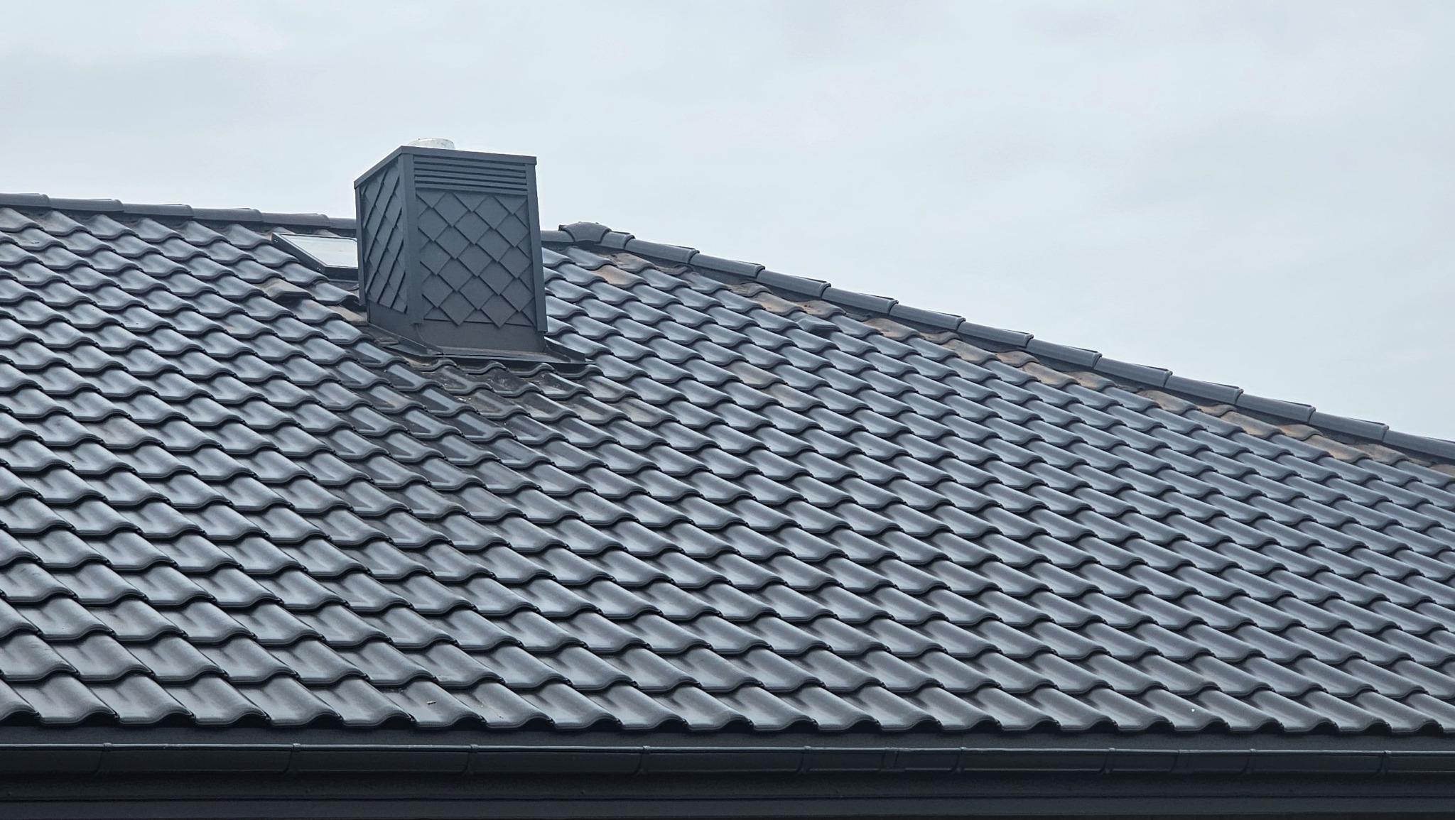 Ciemna dachówka ceramiczna na dachu domu z kominem w zbliżeniu. Widoczne okno dachowe. Dach po renowacji, czysty i zadbany.