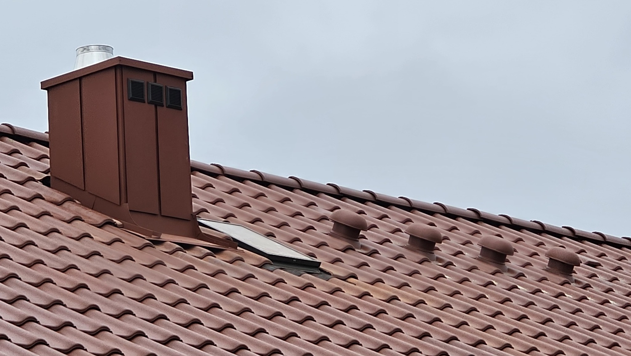 Czerwona dachówka ceramiczna z brązowym kominem i wywietrznikami na tle pochmurnego nieba. Widoczny fragment okna dachowego.
