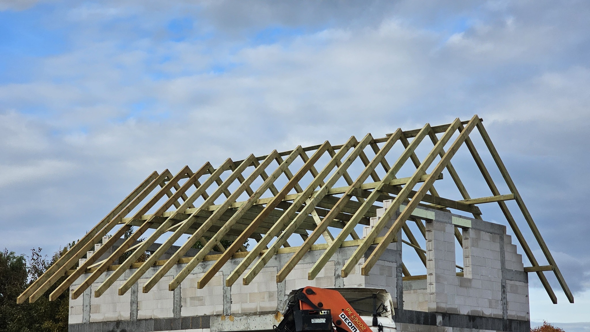Wznoszenie więźby dachowej z jasnego drewna na budynku z białych bloczków, widok z dołu na tle pochmurnego nieba. Fragment żurawia budowlanego w kadrze.