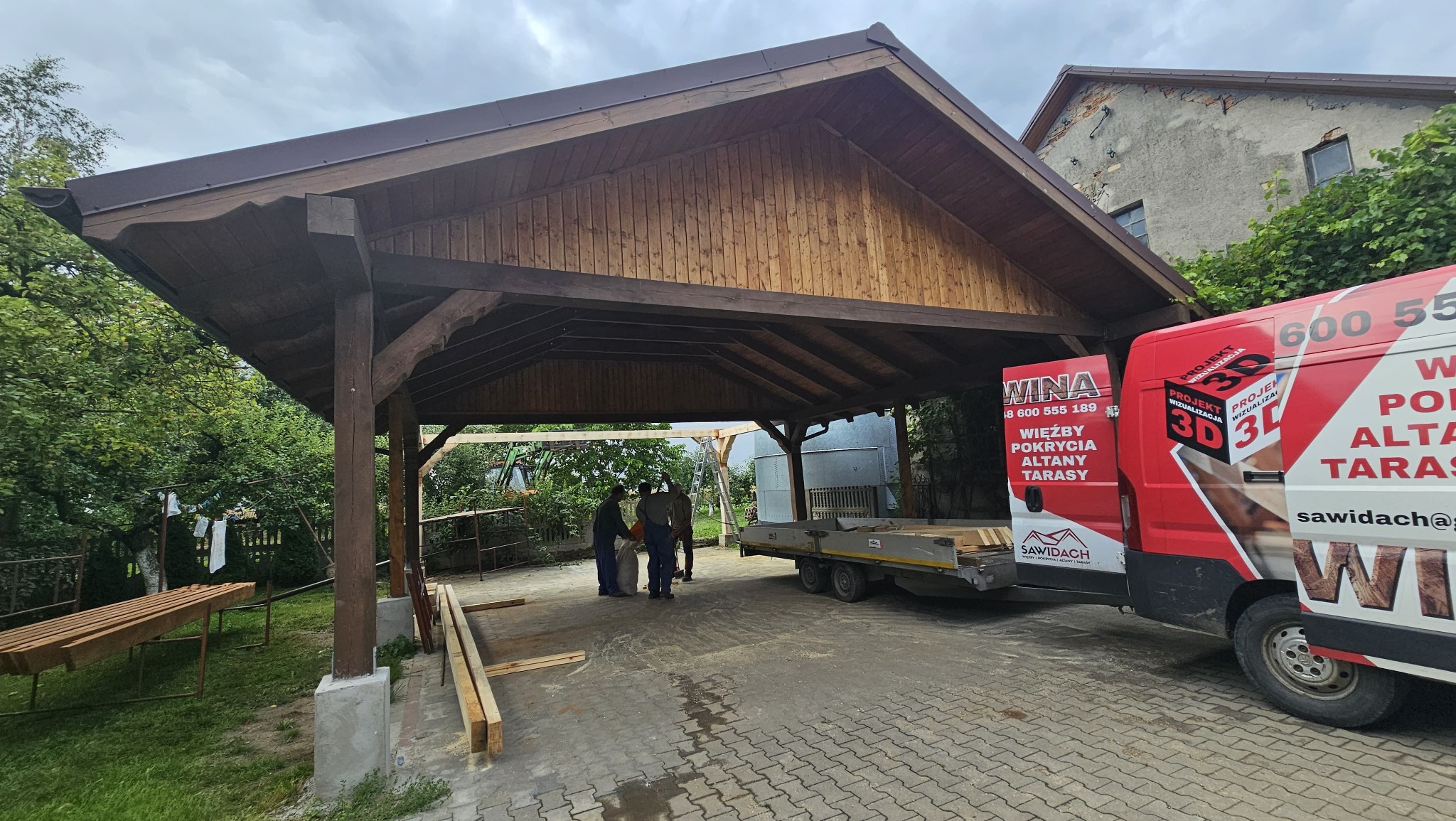 Drewniana wiata z ciemnym dachem w trakcie montażu. Na pierwszym planie samochód dostawczy z reklamą firmy. Widoczne elementy konstrukcyjne i ekipa montażowa.