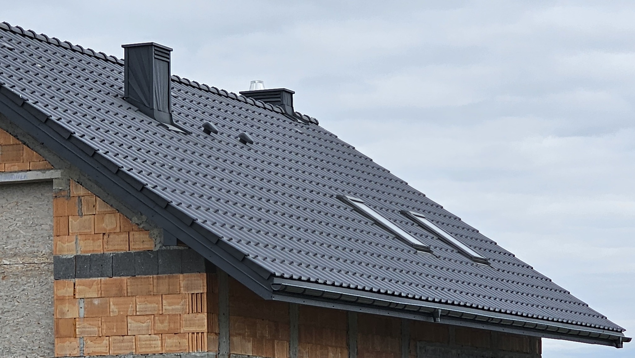 Ciemnoszary dach z dachówki ceramicznej z dwoma oknami dachowymi i kominem na tle pochmurnego nieba, widoczny fragment muru z cegły.