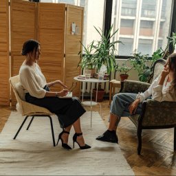 Natalia Szaniawska - Sesja terapeutyczna w jasnym gabinecie: kobieta w eleganckiej bluzce notuje, pacjentka siedzi zamyślona w fotelu, otoczona roślinami i drewnianym parawanem.