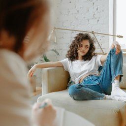 Natalia Szaniawska - Kobieta z kręconymi włosami siedzi na fotelu, ubrana w białą koszulkę i dżinsy, podczas sesji. W tle widoczny biały mur z cegły i okno.