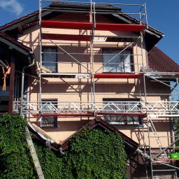 BĄK WOJCIECH USŁUGI REMONTOWE WOJT-MAL - Dom jednorodzinny w trakcie prac elewacyjnych. Rusztowanie obejmuje budynek z oknami zabezpieczonymi folią. Widoczne elementy dachu i zielona roślinność.