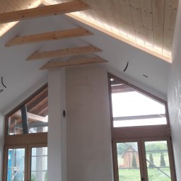 BĄK WOJCIECH USŁUGI REMONTOWE WOJT-MAL - Wnętrze poddasza z drewnianym wykończeniem sufitu i ścian, duże okna w kształcie trójkątów, ściany przygotowane pod malowanie, surowy stan wykończenia.