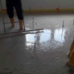 BĄK WOJCIECH USŁUGI REMONTOWE WOJT-MAL - Wylewka samopoziomująca w trakcie rozprowadzania. Widoczne żółte buty robocze i narzędzia. Świeża, mokra powierzchnia podłogi z wystającymi elementami.