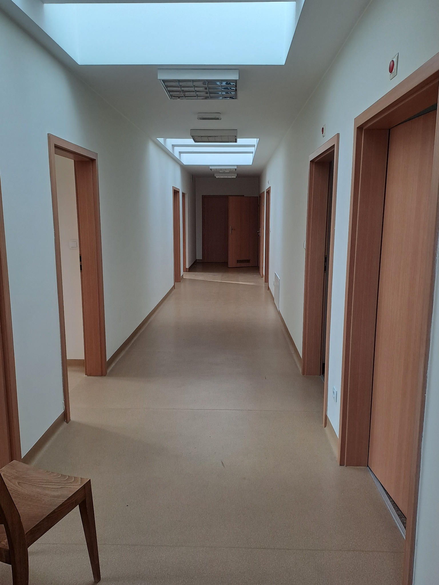 Długi korytarz z jasnymi ścianami, drewnianymi drzwiami i linoleum. Sufit z oknami dachowymi i lampami. Krzesło w rogu kadru. Widok na wnętrze po remoncie.