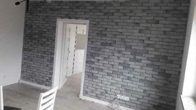 Ściana wykończona szarą cegłą dekoracyjną w pokoju z białymi drzwiami i podłogą. Na podłodze wałek malarski. Widok na wnętrze przez otwarte drzwi.