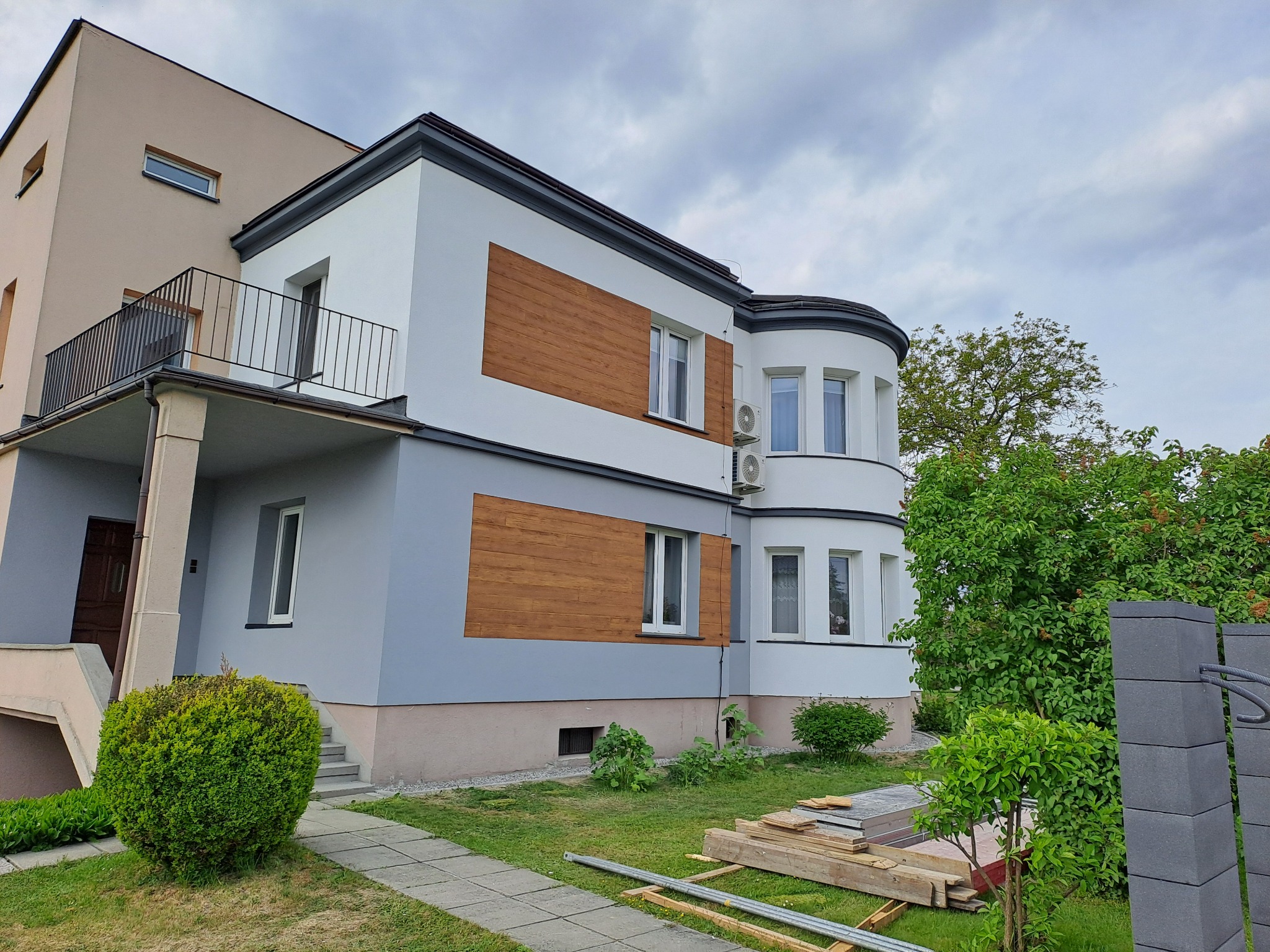 Elewacja domu po renowacji: biało-szara kolorystyka z drewnianymi panelami. Widoczne elementy wykończeniowe i otoczenie budynku. Ujęcie z poziomu ogrodu.