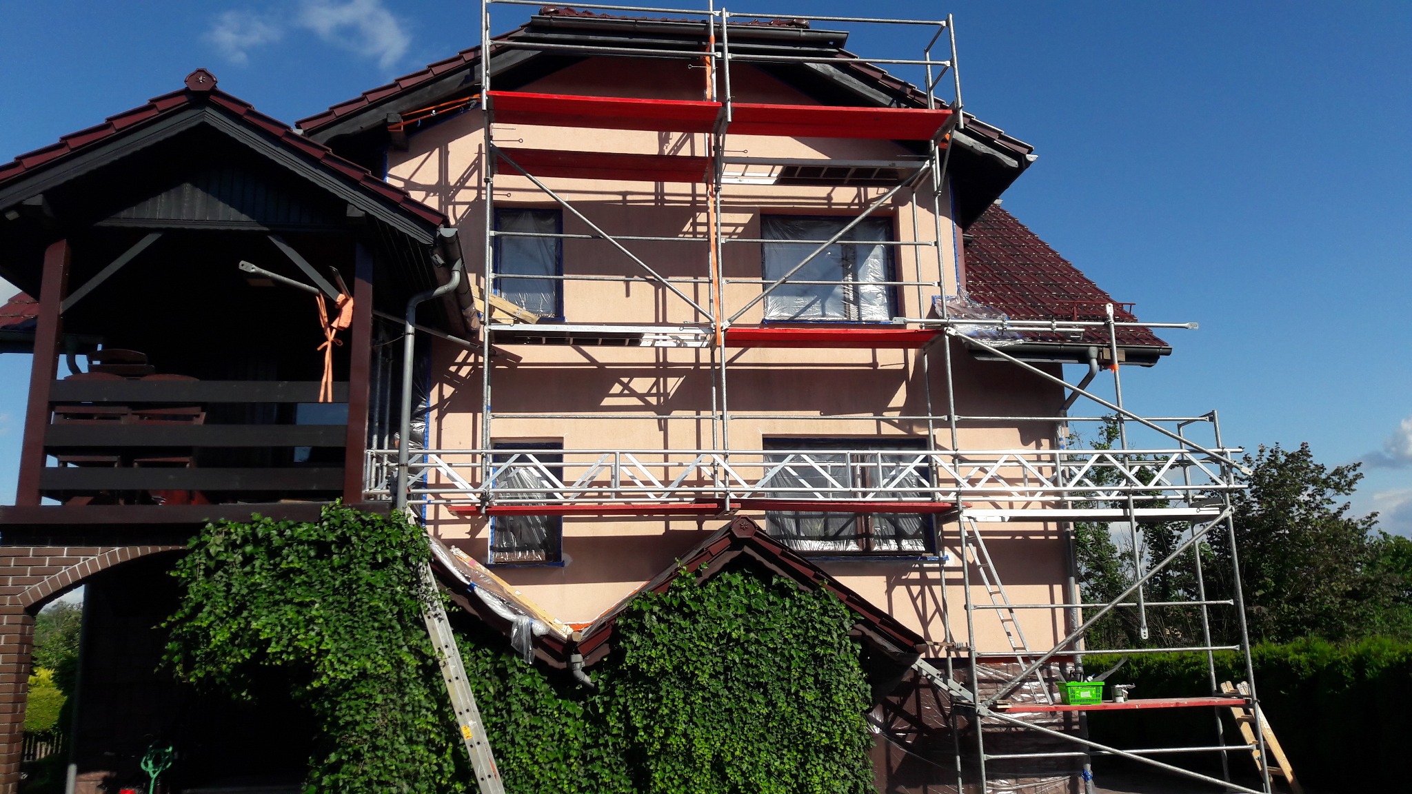 Dom jednorodzinny w trakcie prac elewacyjnych. Rusztowanie obejmuje budynek z oknami zabezpieczonymi folią. Widoczne elementy dachu i zielona roślinność.
