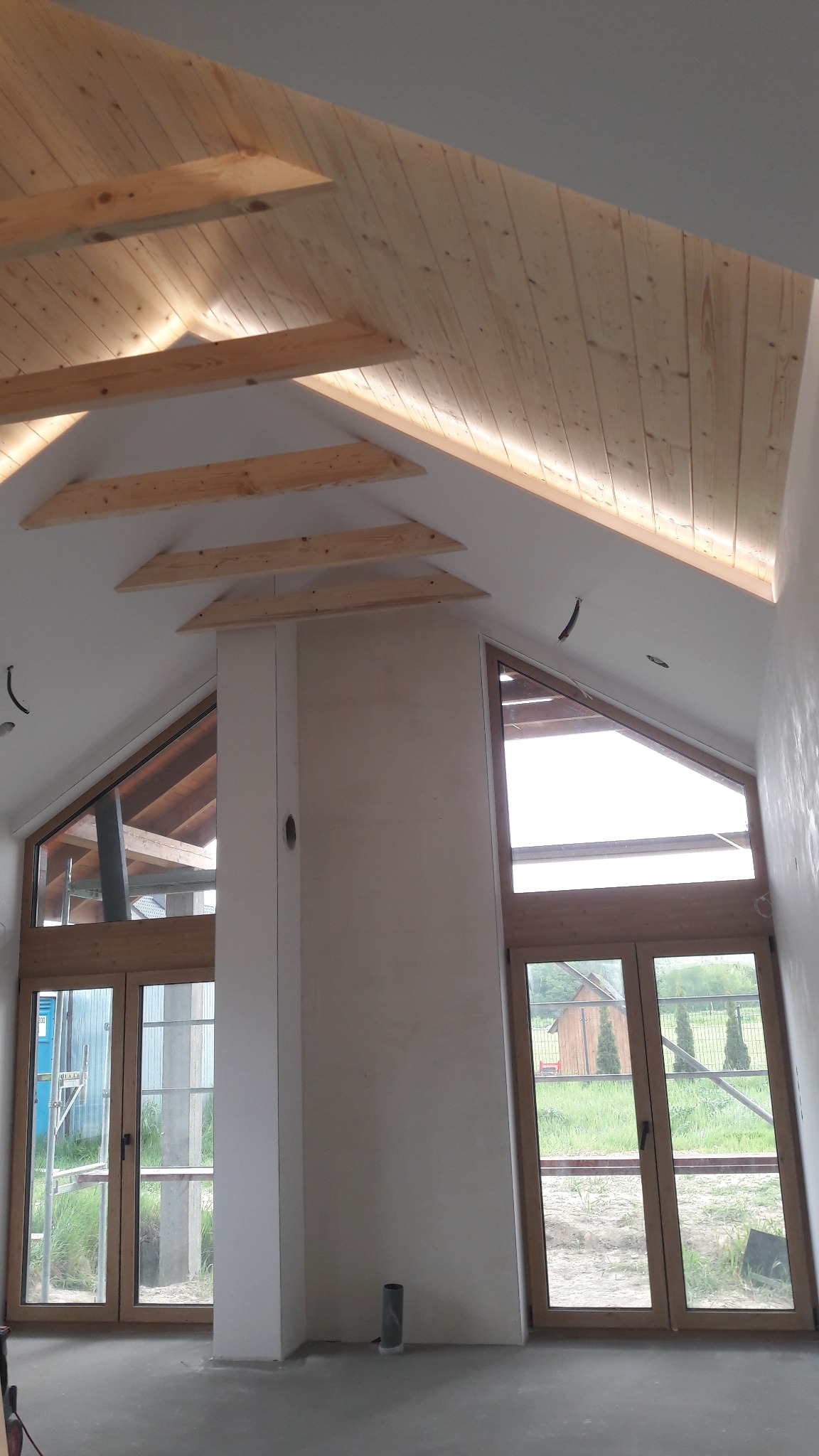 Wnętrze poddasza z drewnianym wykończeniem sufitu i ścian, duże okna w kształcie trójkątów, ściany przygotowane pod malowanie, surowy stan wykończenia.