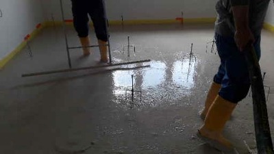 Wylewka samopoziomująca w trakcie rozprowadzania. Widoczne żółte buty robocze i narzędzia. Świeża, mokra powierzchnia podłogi z wystającymi elementami.