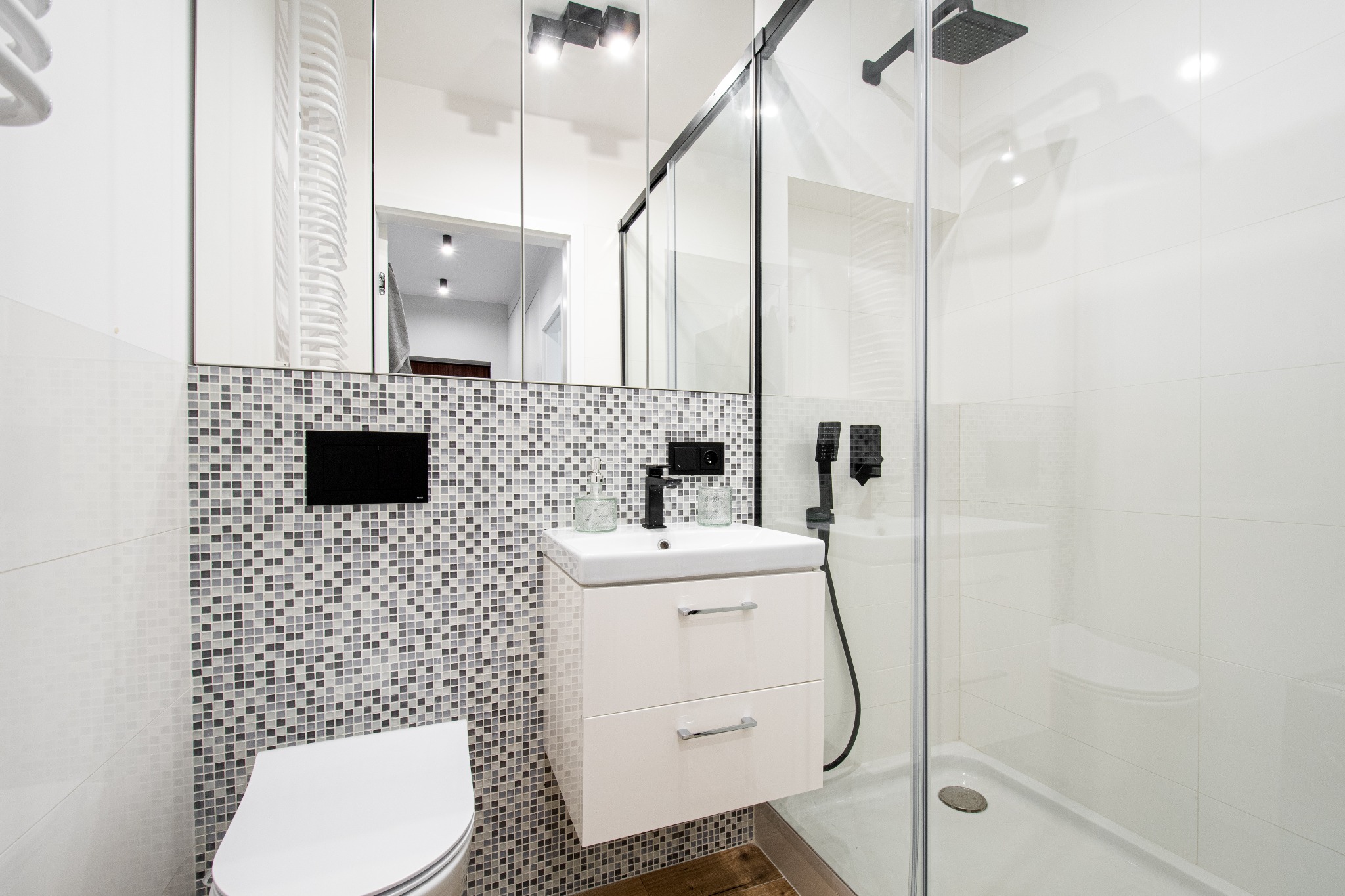 Nowoczesna łazienka z kabiną prysznicową typu walk-in, umywalką na szafce i mozaiką na ścianie. Minimalistyczny design z czarnymi akcentami i jasnymi płytkami.