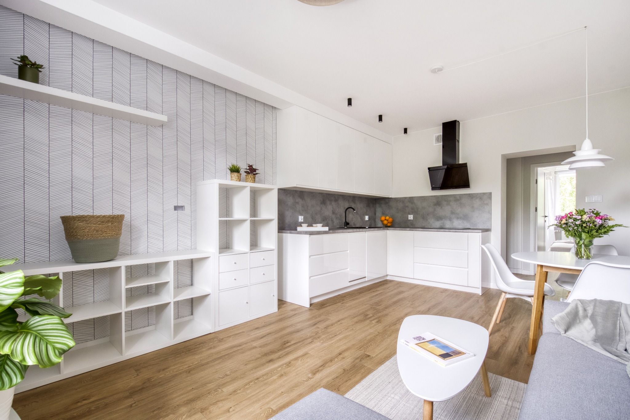 Jasne, przestronne wnętrze: biała zabudowa kuchenna z szarym blatem, drewniana podłoga, wzorzysta tapeta i minimalistyczne meble w salonie. Nowoczesny design.