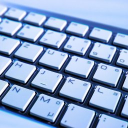 Andrzej Piróg - Klawiatura laptopa z białymi klawiszami podświetlonymi na niebiesko, widok z bliska pod kątem. Widoczne litery i cyfry na klawiszach.