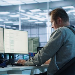 Andrzej Piróg - Programista w szarej bluzie pisze kod na kilku monitorach w nowoczesnym biurze. Widoczne słuchawki na biurku i fragment innego pracownika w tle.