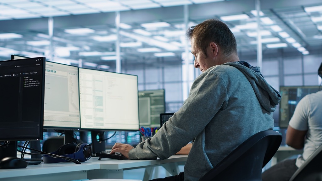Programista w szarej bluzie pisze kod na kilku monitorach w nowoczesnym biurze. Widoczne słuchawki na biurku i fragment innego pracownika w tle.
