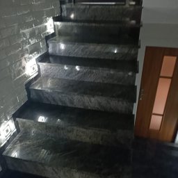 LUKMA MAGDALENA PIASECKA - Nowoczesne schody z ciemnego kamienia z subtelnym oświetleniem LED wzdłuż ściany z dekoracyjną fakturą cegły. Widoczne drzwi w tle.