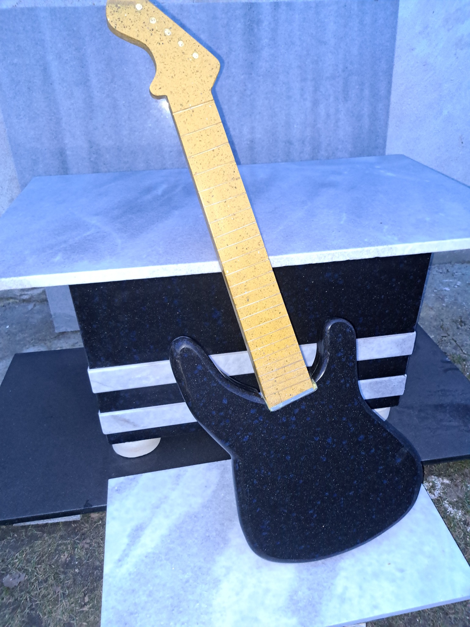 Nietypowy nagrobek w kształcie gitary elektrycznej z ciemnego kamienia, oparty o jasny, kamienny postument. Widoczne detale wykończenia i faktura kamienia.